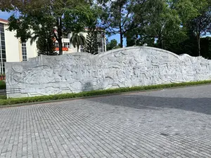 Bức ảnh chụp một không gian công cộng ngoài trời, nổi bật với một bức phù điêu lớn bằng đá. Bức phù điêu này được chạm khắc công phu, mô tả các cảnh sinh hoạt và chiến đấu, có thể là các sự kiện lịch sử. Trên đó có hình ảnh nhiều người, bao gồm các binh lính cầm vũ khí và người dân chèo thuyền (ghe) trên sông. Phía trước bức phù điêu là một khoảng sân rộng được lát đá và một hàng rào cây xanh thấp. Phía sau là nhiều cây xanh và một tòa nhà lớn. Trên tòa nhà có một băng rôn màu đỏ chữ vàng, tuy nhiên chữ bị che khuất một phần và không đọc được toàn bộ, chỉ có thể nhận ra các ký tự như '...NGHĨA VIỆ...' và '...ƠN NĂM...'.。（日本語訳）。写真は屋外の公共空間を撮影したもので、大きな石のレリーフが際立っています。このレリーフは精巧に彫られており、生活や戦闘の場面を描写しており、歴史的な出来事である可能性があります。そこには多くの人々の姿があり、武器を持った兵士や川で船（小舟）を漕ぐ人々が含まれています。レリーフの前には石畳の広い中庭と低い生け垣があります。後ろには多くの緑の木々と大きな建物があります。建物には金色の文字が入った赤い横断幕がありますが、文字の一部が隠れていて全部を読むことはできず、「...NGHĨA VIỆ...」や「...ƠN NĂM...」のような文字しか認識できません。