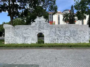 Bức ảnh chụp một bức tường phù điêu lớn bằng đá, nằm trong một không gian công cộng như công viên hoặc quảng trường tại Thanh Hóa. Bức tường có một cổng vòm ở trung tâm và được chạm khắc tinh xảo các hoạt cảnh lịch sử. Các hình khắc mô tả các đoàn quân, binh lính, một số đang cưỡi ngựa trong trận chiến, và cả những người dân. Phía trước bức phù điêu là một hàng cây bụi được cắt tỉa và một khoảng sân rộng lát đá. Ở phía sau, có nhiều cây xanh và một tòa nhà lớn, hiện đại với mái vòm. Trên tòa nhà có treo cờ Việt Nam và các tấm pa-nô màu đỏ, trong đó có một tấm mang biểu tượng búa liềm và một tấm khác có hình hoa sen.。（日本語訳）。写真には、タインホア省の公園や広場のような公共スペースにある、大きな石のレリーフの壁が写っている。壁の中央にはアーチ門があり、歴史的な場面が精巧に彫刻されている。彫刻は、軍隊、兵士、その中には戦場で馬に乗っている者、そして一般の人々を描写している。レリーフの前には、刈り込まれた植え込みの列と、石畳の広い中庭がある。後方には、多くの緑の木々と、ドーム屋根を持つ大きな近代的な建物がある。建物にはベトナム国旗と赤いパネルが掲げられており、その中にはハンマーと鎌のシンボルが描かれたものと、蓮の花の絵が描かれたものがある。