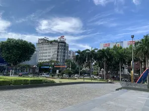 Bức ảnh chụp một khung cảnh đô thị tại một quảng trường hoặc ngã tư lớn ở thành phố Thanh Hóa vào một ngày nắng đẹp, trời xanh và có mây trắng. Nổi bật trong ảnh là một tòa nhà cao tầng, trên nóc có các biển hiệu màu đỏ ghi 'HOTEL MỊ CHUNG' và một biển hiệu khác ghi 'Coffee'. Ở phía xa bên phải, có một tấm biển quảng cáo lớn với dòng chữ '3 TINH HOA VỊ ĐẬM CHẤT' và logo bia '333'. Phía trước các tòa nhà là một con đường có ô tô và xe máy đang lưu thông. Giữa quảng trường và con đường là một khu vực công viên trồng nhiều cây xanh, cây cọ và các hàng rào cây được cắt tỉa. Sân quảng trường rộng lớn được lát gạch. Có một vài người đang di chuyển trên đường phố.。（日本語訳）。写真は、青空に白い雲が浮かぶ晴れた日の、タインホア市にある大きな広場または交差点の都市風景を捉えたものです。写真の中で目立っているのは高層ビルで、その屋上には「HOTEL MỊ CHUNG」と書かれた赤い看板と、「Coffee」と書かれた別の看板があります。右奥には、「3 TINH HOA VỊ ĐẬM CHẤT」という文字と「333」ビールのロゴが入った大きな広告看板があります。建物の前には、自動車やバイクが通行している道路があります。広場と道路の間には、多くの緑の木々、ヤシの木、そして刈り込まれた生け垣が植えられた公園エリアがあります。広大な広場の地面はタイルで舗装されています。通りには数人の人が移動しています。