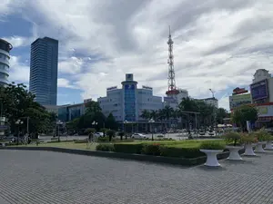 Bức ảnh chụp một khung cảnh đô thị hiện đại vào ban ngày tại một quảng trường hoặc vòng xoay lớn, có khả năng là ở thành phố Thanh Hóa. Bầu trời có mây nhưng thời tiết quang đãng. Ở phía trước là một khoảng sân rộng lát gạch và một khu vườn công cộng được cắt tỉa gọn gàng với hàng rào cây xanh, bãi cỏ và những chậu cây cảnh lớn màu trắng. Phía sau là các con đường với nhiều phương tiện giao thông như ô tô và xe máy đang di chuyển. Có nhiều tòa nhà cao tầng hiện đại, nổi bật là tòa nhà màu xanh của VNPT với logo "VNPT" và chữ "vinaphone". Bên trái là một tòa nhà chọc trời bằng kính. Ở trung tâm có một cột tháp viễn thông cao sơn hai màu đỏ và trắng. Các biển hiệu có thể nhận dạng bao gồm: "VNPT", "vinaphone", "18001166" trên cột tháp, "LONG ANH HOTEL", "BACER CITY TRƯỜNG ĐUA", "HAYSPO", "TOP 10", và "CÔNG TY CỔ PHẦN". Hoạt động chính là giao thông đô thị và sinh hoạt thường ngày của người dân.。（日本語訳）。写真は、おそらくタインホア市にある大きな広場またはロータリーで、日中の現代的な都市の風景を撮影したものです。空は曇っていますが、天気は晴れやかです。手前には、広くタイルが敷かれた広場と、緑の生け垣、芝生、そして大きな白い観葉植物の鉢がある、きれいに手入れされた公園があります。後方には道路があり、自動車やバイクなど多くの交通手段が移動しています。多くの近代的な高層ビルがあり、目立つのは「VNPT」のロゴと「vinaphone」の文字があるVNPTの青いビルです。左側にはガラス張りの超高層ビルがあります。中央には、赤と白の2色で塗装された高い通信塔があります。認識できる看板には、塔にある「VNPT」、「vinaphone」、「18001166」、「LONG ANH HOTEL」、「BACER CITY TRƯỜNG ĐUA」、「HAYSPO」、「TOP 10」、そして「CÔNG TY CỔ PHẦN」が含まれます。主な活動は、都市の交通と人々の日常生活です。