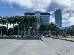 Bức ảnh chụp một quảng trường công cộng rộng lớn tại trung tâm thành phố Thanh Hóa vào một ngày trời trong xanh, có mây. Phía trước là một khoảng sân rộng được lát đá xám. Nổi bật trong khung cảnh là các tòa nhà hiện đại. Một tòa nhà có kiến trúc cong với logo 'BIDV' trên nóc. Phía sau nó là một tòa nhà chọc trời cao tầng bằng kính. Một tòa nhà khác ở phía xa bên phải có biển hiệu 'XINGFA'. Ngoài ra còn có biển hiệu của 'BSC' (Công ty Cổ phần Chứng khoán BIDV) trên tòa nhà BIDV. Dọc theo quảng trường và đường phố là hàng cây cọ cao và nhiều cây xanh khác. Trên đường phố, có nhiều ô tô và xe máy đang di chuyển và đậu đỗ, thể hiện hoạt động giao thông thường ngày của đô thị. Một vài người đang ngồi trên ghế đá công cộng.。（日本語訳）。この写真は、晴れて雲のある日にタインホア市の中心部にある広大な公共広場を撮影したものです。手前には、灰色の石で舗装された広い広場があります。風景の中で際立っているのは、近代的な建物です。ある建物は湾曲した建築様式で、屋上には「BIDV」のロゴがあります。その後ろには、ガラス張りの高層ビルがあります。右奥にある別の建物には、「XINGFA」の看板があります。さらに、BIDVの建物には「BSC」（BIDV証券株式会社）の看板もあります。広場と通りに沿って、高いヤシの木や他の多くの緑の木々が並んでいます。通りには、多くの車やバイクが移動したり駐車したりしており、都市の日常的な交通活動を示しています。何人かの人々が公共の石のベンチに座っています。