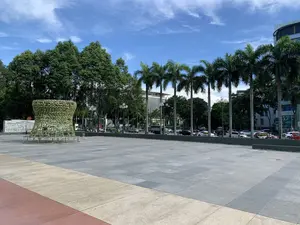 Bức ảnh chụp một quảng trường công cộng rộng lớn vào một ngày nắng đẹp. Nền quảng trường được lát gạch đá với các màu xám, be và nâu đỏ. Ở phía bên trái của quảng trường có một công trình trang trí lớn, có hình dạng giống một cái trống hoặc một chiếc bình hoa, được phủ kín bằng hoa giả màu xanh lá cây và trắng. Phía sau công trình trang trí này là một bức phù điêu bằng đá.。Xa hơn một chút là một con đường có xe cộ qua lại, bao gồm ô tô và xe máy. Dọc theo con đường là một hàng cây cọ cao và các loại cây xanh khác, cùng với các cột đèn đường. Trong hậu cảnh là các tòa nhà cao tầng hiện đại. Bầu trời trong xanh với vài gợn mây trắng. Có thể nhìn thấy một số biển hiệu trên các tòa nhà, nhưng chữ viết quá nhỏ và mờ để đọc được, tuy nhiên có thể nhận ra logo của ngân hàng BIDV.。（日本語訳）。写真は、晴れた日の広大な公共広場を写している。広場の地面は、灰色、ベージュ、赤褐色の石タイルで舗装されている。広場の左側には、太鼓または花瓶のような形をした大きな装飾物があり、緑と白の造花で覆われている。この装飾物の後ろには、石のレリーフがある。少し奥には、自動車やバイクなどの車両が往来する道路がある。道路に沿って、高いヤシの木やその他の緑樹の列があり、街灯も並んでいる。背景には、近代的な高層ビル群がある。空は青く澄んでおり、白い雲がいくつか浮かんでいる。しかし、BIDV銀行のロゴは認識できる。