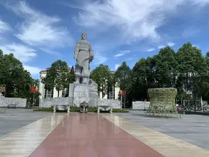 Bức ảnh chụp một quảng trường rộng lớn vào một ngày trời trong xanh, có nắng và mây trắng. Trung tâm của quảng trường là một tượng đài lớn bằng đá, tạc hình Anh hùng dân tộc Lê Lợi trong trang phục cổ, tư thế oai nghiêm. Trên bệ tượng có khắc các dòng chữ: 'ANH HÙNG DÂN TỘC', 'LÊ LỢI', và '1385 - 1433'. Phía trước tượng đài có một lư hương lớn bằng đá và hai bên là hai băng ghế đá được chạm khắc. Sân quảng trường được lát đá với một lối đi chính màu đỏ sẫm dẫn đến chân tượng đài. Xung quanh tượng đài là cây cối xanh tươi, và phía sau là một tòa nhà lớn. Ở hai bên tượng đài có các bức tường phù điêu bằng đá. Ở phía bên trái có thể thấy cờ đỏ sao vàng (Quốc kỳ Việt Nam) và cờ búa liềm (cờ Đảng). Khung cảnh chung yên tĩnh, trang nghiêm.。（日本語訳）。青空が広がり、太陽が照り、白い雲が浮かぶ日の、広大な広場を写した写真です。広場の中央には、民族の英雄レ・ロイが古来の衣装をまとい、威厳のある姿で彫られた大きな石の像があります。像の台座には、「民族の英雄」、「レ・ロイ」、そして「1385 - 1433」という文字が刻まれています。像の前には大きな石の香炉があり、両側には彫刻が施された2つの石のベンチがあります。広場の地面は石で舗装されており、濃い赤色の主要な通路が像の足元まで続いています。像の周りには青々とした木々があり、その後ろには大きな建物があります。像の両側には石の浮き彫りの壁があります。左側には、金星紅旗（ベトナム国旗）とハンマーと鎌の旗（党旗）が見えます。全体的な光景は静かで、荘厳です。