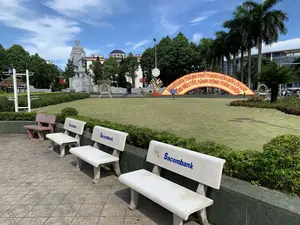 Bức ảnh chụp một không gian công cộng, dường như là một công viên hoặc quảng trường tại thành phố Thanh Hóa vào một ngày nắng đẹp, trời trong xanh. Ở phía trước là một hàng ghế đá xi măng đặt trên vỉa hè lát gạch, trong đó có ba chiếc ghế màu trắng có in chữ 'Sacombank' màu xanh. Phía sau hàng ghế là một bãi cỏ xanh rộng, được cắt tỉa gọn gàng. Trên bãi cỏ có một tấm biển nhỏ màu xanh ghi 'KHÔNG DẪM LÊN CỎ'. Ở giữa công viên, có một cổng chào lớn hình vòng cung được trang trí bằng hoa màu vàng với dòng chữ 'NGƯỜI DÂN THÀNH PHỐ THANH HÓA NÓI LỜI HAY, LÀM VIỆC TỐT, HÀNH ĐỘNG THÂN THIỆN'. Phía sau bãi cỏ là tượng đài Anh hùng dân tộc Lê Lợi bằng đá, trên bệ tượng có khắc chữ 'ANH HÙNG DÂN TỘC LÊ LỢI'. Xung quanh khu vực có nhiều cây xanh, bao gồm cả những cây cọ cao, và phía xa là các tòa nhà.。（日本語訳）。写真は、晴れた美しい日、青空が広がるタインホア市の公園か広場と思われる公共スペースを写しています。手前にはタイル張りの歩道にセメント製の石のベンチが並んでおり、その中には青い文字で「Sacombank」と印字された白いベンチが3つあります。ベンチの後ろには、きれいに刈り込まれた広大な緑の芝生が広がっています。芝生の上には、「KHÔNG DẪM LÊN CỎ」（芝生に入らないでください）と書かれた小さな青い看板があります。公園の中央には、黄色い花で飾られた大きなアーチ型の歓迎ゲートがあり、「NGƯỜI DÂN THÀNH PHỐ THANH HÓA NÓI LỜI HAY, LÀM VIỆC TỐT, HÀNH ĐỘNG THÂN THIỆN」（タインホア市民は、良い言葉を話し、良い行いをし、親しみやすく行動する）という文字が書かれています。芝生の後ろには、石造りの民族英雄レ・ロイの像があり、台座には「ANH HÙNG DÂN TỘC LÊ LỢI」（民族英雄レ・ロイ）という文字が刻まれています。周辺には、高いヤシの木を含む多くの緑の木々があり、遠くには建物が見えます。