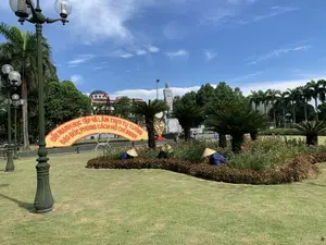 Bức ảnh chụp một công viên công cộng ở Thanh Hóa vào một ngày nắng đẹp. Ở trung tâm là một bãi cỏ xanh rộng lớn với các bồn hoa được chăm sóc cẩn thận. Có hai hoặc ba công nhân đang làm vườn, đội nón lá truyền thống, cúi xuống chăm sóc cây trong bồn hoa. Phía sau họ là một cổng chào lớn hình vòng cung màu vàng với dòng chữ đỏ: 'ĐẨY MẠNH HỌC TẬP VÀ LÀM THEO TƯ TƯỞNG, ĐẠO ĐỨC, PHONG CÁCH HỒ CHÍ MINH'. Ở phía xa hơn là Tượng đài Lê Lợi bằng đá, đứng trên một bệ lớn. Phía sau tượng đài là một tòa nhà kiến trúc hiện đại có mái vòm và lá cờ Việt Nam. Khung cảnh được bao quanh bởi cây xanh, bao gồm cả cây cọ, và các cột đèn trang trí kiểu cổ. Bầu trời trong xanh với vài gợn mây trắng.。（日本語訳）。晴れた日にタインホア省のある公共公園を撮影した写真です。中央には、手入れの行き届いた花壇のある広大な緑の芝生があります。2、3人の作業員が庭仕事をしており、伝統的なノンラーをかぶり、かがんで花壇の植物の手入れをしています。彼らの後ろには、赤い文字で「ホーチミンの思想、道徳、スタイルに学び、従うことを推進する」と書かれた大きな黄色いアーチ型の門があります。さらに奥には、大きな台座の上に立つ石造りのレ・ロイ像があります。像の後ろには、ドーム屋根とベトナム国旗のある近代的な建築の建物があります。風景は、ヤシの木を含む緑の木々や、アンティーク調の装飾的な街灯に囲まれています。空は青く澄んでおり、白い雲がいくつか浮かんでいます。