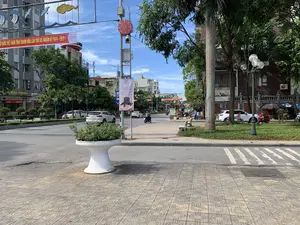 Các dòng chữ ghi nhận được bao gồm một băng rôn đỏ lớn treo ngang đường với nội dung 'ĐẠI HỘI MẶT TRẬN TỔ QUỐC VIỆT NAM TỈNH THANH HÓA LẦN THỨ XV, NHIỆM KỲ 2024 - 2029。' và logo 'Viettel'. Một phướn quảng cáo dọc cho sự kiện khai trương ('Grand OPENING') của cửa hàng 'NHAT LEATHER' vào ngày 05.07.2024 tại Phường Điện Biên, Thành phố Thanh Hóa. Ngoài ra còn có các biển hiệu cửa hàng như 'KIM CHUNG CÔNG TY VÀNG...'. Khung cảnh là một con đường đô thị tại thành phố Thanh Hóa vào ban ngày, dưới trời nắng. Con đường có nhiều ô tô và xe máy đang lưu thông. Hai bên đường là các tòa nhà cao tầng, cây xanh và vỉa hè rộng rãi được lát gạch. Trên vỉa hè có người dân ngồi trên ghế đá và một số quán hàng rong nhỏ có ô che. Một chậu cây cảnh lớn màu trắng được đặt trên vỉa hè ở phía trước. Hoạt động chính là giao thông và sinh hoạt thường ngày của người dân thành phố.。（日本語訳）。確認された文字には、道路を横切って掲げられた「第15回タインホア省ベトナム祖国戦線大会、任期2024年～2029年。」という内容の大きな赤い横断幕と、「Viettel」のロゴが含まれます。2024年7月5日にタインホア市ディエンビエン区で行われる「NHAT LEATHER」店のグランドオープニングイベントのための縦長の広告旗が一つあります。その他にも、「KIM CHUNG 金会社…」のような店舗の看板があります。風景は、晴天の日中におけるタインホア市の都市の道路です。道路には多くの自動車とバイクが通行しています。道路の両側には高層ビル、緑の木々、そしてタイル張りの広い歩道があります。歩道の上には、石のベンチに座っている人々や、日傘をさしたいくつかの小さな露店があります。手前の歩道には、大きな白い観葉植物の鉢が一つ置かれています。主な活動は、交通と都市の人々の日常生活です。