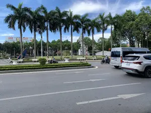 Bối cảnh là một con đường nhựa tại một khu vực đô thị, có thể là một vòng xuyến hoặc ngã tư, vào một ngày nắng đẹp với bầu trời xanh và mây trắng. Bên đường là một quảng trường hoặc công viên công cộng. Trong công viên, có một tượng đài lớn bằng đá màu trắng, khắc họa một nhân vật lịch sử. Một hàng cây cọ cao và thẳng tắp được trồng dọc theo quảng trường. Ngoài ra còn có các bãi cỏ xanh, hàng rào cây được cắt tỉa gọn gàng, và những chiếc ghế đá màu trắng. Phía xa có một tấm biển quảng cáo lớn của hãng bia 333 với dòng chữ '3 TINH HOA VỊ ĐẬM CHẤT'. Trên đường, có các phương tiện đang tham gia giao thông, bao gồm một chiếc ô tô SUV màu trắng, một chiếc xe khách nhỏ màu bạc có chữ 'LUXURY' trên cửa sổ, và hai người đang đi xe máy. Mặt đường có kẻ vạch phân làn và mũi tên chỉ hướng.。（日本語訳）。背景は都市部のアスファルト道路で、ロータリー交差点または十字路の可能性があります。青い空と白い雲が広がる晴れた日の光景です。道端には広場または公共の公園があります。公園の中には、歴史上の人物を描いた大きな白い石の像があります。広場に沿って、高くてまっすぐなヤシの木が一列に植えられています。その他にも、緑の芝生、きれいに刈り込まれた生垣、そして白い石のベンチがあります。遠くには、「3 TINH HOA VỊ ĐẬM CHẤT」という文字が書かれた333ビールの大きな広告看板があります。道路では、白いSUV車、窓に「LUXURY」と書かれた銀色の小型バス、そしてバイクに乗った2人を含む車両が通行しています。路面には車線区分線と方向指示の矢印が引かれています。