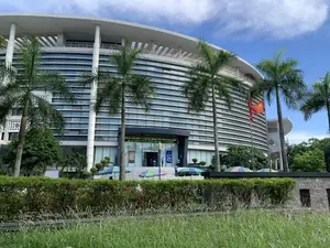 Bức ảnh chụp mặt tiền của một tòa nhà công vụ lớn, hiện đại có kiến trúc hình vòng cung dưới bầu trời xanh. Trên biển hiệu chính phía trên lối vào có dòng chữ: 'TRUNG TÂM PHỤC VỤ HÀNH CHÍNH CÔNG TỈNH THANH HÓA' và địa chỉ 'ĐỊA CHỈ: 28 ĐẠI LỘ LÊ LỢI - PHƯỜNG ĐIỆN BIÊN - TP. THANH HÓA'. Đây là Trung tâm Phục vụ Hành chính công của tỉnh Thanh Hóa. Phía trước tòa nhà có trồng nhiều cây cọ cao. Bên phải lối vào có một cột cờ treo lá cờ đỏ sao vàng của Việt Nam. Một vài người dân và nhân viên đang di chuyển ở khu vực bậc thang và cửa ra vào. Gần lối vào có một vài chiếc ô lớn, nhiều màu sắc được dựng lên, trên một chiếc ô có ghi số điện thoại '02373 888 999'. Phía trước, ngăn cách bởi một hàng rào cây xanh, là một khu vực cỏ mọc cao.。（日本語訳）。青空の下、弧を描くような建築様式の、大きくて近代的な公共サービスの建物の正面を写した写真です。入り口の上にあるメインの看板には、「TRUNG TÂM PHỤC VỤ HÀNH CHÍNH CÔNG TỈNH THANH HÓA」という文字と、「ĐỊA CHỈ: 28 ĐẠI LỘ LÊ LỢI - PHƯỜNG ĐIỆN BIÊN - TP. THANH HÓA」という住所が書かれています。これはタインホア省の公共行政サービスセンターです。建物の前には、背の高いヤシの木がたくさん植えられています。入り口の右側には、ベトナムの金星紅旗を掲げた旗竿があります。階段や出入り口のあたりで、数人の市民や職員が移動しています。入り口の近くには、いくつかのカラフルで大きな傘が立てられており、そのうちの一つの傘には「02373 888 999」という電話番号が書かれています。手前には、生け垣で隔てられた、草丈の高い草地があります。