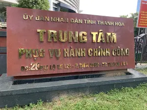 Bức ảnh chụp một tấm biển hiệu lớn bằng đá màu nâu đỏ, đặt trên một bệ đá màu đen bên cạnh bãi cỏ. Đây là biển hiệu của một cơ quan hành chính công. Toàn bộ chữ viết trên biển là chữ nổi màu vàng đồng, được ghi lại như sau:。- Dòng trên cùng: "ỦY BAN NHÂN DÂN TỈNH THANH HÓA"。- Dòng chữ lớn ở giữa: "TRUNG TÂM PHỤC VỤ HÀNH CHÍNH CÔNG"。- Dòng địa chỉ ở dưới: "ĐC: 28 ĐẠI LỘ LÊ LỢI - P. ĐIỆN BIÊN - TP. THANH HÓA"。Dựa trên nội dung, đây là Trung tâm Phục vụ Hành chính công tỉnh Thanh Hóa. Phía sau biển hiệu là một tòa nhà hiện đại, cây xanh và cổng vào. Ở phía bên phải, có một biển báo thông tin màu đỏ khác nhưng chỉ đọc được một phần.。（日本語訳）。写真は、芝生の隣にある黒い石の台座の上に置かれた、赤褐色の石でできた大きな看板を写しています。これは行政機関の看板です。看板の文字はすべてブロンズ色の浮き彫り文字で、次のように書かれています。- 最上段：「タインホア省人民委員会」。- 中央の大きな文字：「行政サービスセンター」。- 下段の住所：「住所：タインホア市ディエンビエン坊レロイ大通り28番地」。内容から、これはタインホア省行政サービスセンターです。看板の後ろには、近代的な建物、緑の木々、そして入口の門があります。右側には、別の赤い案内看板がありますが、一部しか読み取れません。