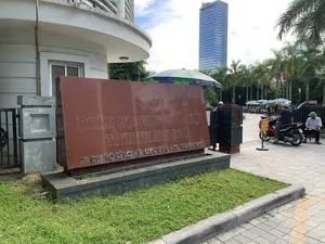 Chữ khắc trên bia đá: 'ĐOÀN ĐẠI BIỂU QUỐC HỘI TỈNH THANH HOÁ', và địa chỉ '28 ĐẠI LỘ LÊ LỢI - P. ĐIỆN BIÊN - TP. THANH HOÁ'. Ngoài ra còn có biển báo nhỏ ghi 'VUI LÒNG DỪNG XE LẤY VÉ VÀO CỔNG'.。Đây là quang cảnh bên ngoài lối vào một tòa nhà văn phòng, cụ thể là trụ sở của Đoàn Đại biểu Quốc hội tỉnh Thanh Hóa. Một tấm bia đá lớn màu nâu đỏ khắc tên cơ quan và địa chỉ được đặt nổi bật ở phía trước. Phía sau tấm bia là khu vực cổng vào và bãi gửi xe máy. Có một vài người đang ở khu vực này; một người đàn ông đội nón cối đang ngồi trên ghế, có thể là nhân viên bảo vệ, và một người khác đội mũ bảo hiểm đang ngồi trên xe máy. Nhiều chiếc ô lớn được dựng lên để che nắng. Ở phía xa, có thể thấy một tòa nhà cao tầng hiện đại bằng kính và hàng cây cọ.。（日本語訳）。石碑に刻まれた文字：「タインホア省国会代表団」、および住所「タインホア市ディエンビエン坊レロイ大通り28番地」。その他に「ゲートに入る際は駐車してチケットをお取りください」と書かれた小さな看板もある。これはオフィスビルの入り口の外の光景で、具体的にはタインホア省国会代表団の本部である。石碑の後ろはゲートとバイク駐車場である。このエリアには数人の人がいる。兵士帽をかぶった男性が椅子に座っており、警備員かもしれない。そしてヘルメットをかぶった別の人がバイクに座っている。日よけのために大きなパラソルがいくつか立てられている。