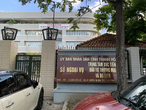 Bức ảnh chụp quang cảnh bên ngoài một tòa nhà văn phòng của cơ quan nhà nước tại thành phố Thanh Hóa. Phía trước tòa nhà có một tấm biển lớn bằng đá màu nâu, khắc chữ vàng ghi rõ thông tin: 'ỦY BAN NHÂN DÂN TỈNH THANH HÓA', 'SỞ NGOẠI VỤ', 'TRUNG TÂM XÚC TIẾN ĐẦU TƯ THƯƠNG MẠI VÀ DU LỊCH', và địa chỉ 'ĐỊA CHỈ: 2 ĐẠI LỘ LÊ LỢI, P. ĐIỆN BIÊN, TP. THANH HÓA'.。Trên mặt tiền của tòa nhà chính, có treo một băng rôn lớn màu trắng với nội dung bằng cả tiếng Việt và tiếng Anh: 'TRUNG TÂM XÚC TIẾN ĐẦU TƯ, THƯƠNG MẠI VÀ DU LỊCH TỈNH THANH HÓA' (THANH HOA INVESTMENT, TRADE AND TOURISM PROMOTION AGENCY) cùng với số điện thoại.。Đây là một tòa nhà hành chính công, có kiến trúc hiện đại nhiều tầng với mặt tiền cong. Khung cảnh cho thấy một ngày trời nắng, có cây xanh bao quanh. Trước cổng có hai chiếc xe ô tô đang đỗ, một chiếc màu trắng và một phần của chiếc màu đỏ. Lối vào có cổng sắt và các cột trụ ốp gạch vuông vắn. Không có hoạt động của con người nào được ghi nhận, chỉ là một cảnh quan tĩnh tại khu vực văn phòng.。（日本語訳）。タインホア市にある国家機関のオフィスビルの外観を撮影した写真です。建物の前には、茶色の石でできた大きな看板があり、金色の文字で「タインホア省人民委員会」、「外務局」、「投資・貿易・観光促進センター」、そして住所「住所：タインホア市ディエンビエン区レロイ大通り2番地」という情報が明記されています。主要な建物の正面には、ベトナム語と英語の両方で「タインホア省投資・貿易・観光促進センター」（THANH HOA INVESTMENT, TRADE AND TOURISM PROMOTION AGENCY）という内容と電話番号が書かれた、白い大きな横断幕が掲げられています。これは公共の行政庁舎で、正面が湾曲した現代的な多層階建ての建築様式です。風景は晴れた日であることを示しており、周囲は緑の木々に囲まれています。門の前には2台の車が駐車しており、1台は白色で、もう1台は赤色の一部が見えます。入り口には鉄の門と、四角いタイルで覆われた柱があります。人の活動は見られず、オフィスエリアの静かな風景が広がっているだけです。