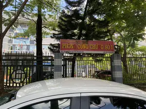 Bức ảnh chụp cổng vào của một cơ quan công quyền, cụ thể là Phòng Công chứng số 1 tại thành phố Thanh Hóa. Có một tấm biển lớn màu đỏ với chữ màu vàng ghi rõ các thông tin sau:。- Dòng trên cùng: "SỞ TƯ PHÁP THANH HÓA"。- Dòng chữ lớn ở giữa: "PHÒNG CÔNG CHỨNG SỐ 1"。- Dòng dưới cùng: "ĐC: 32 ĐẠI LỘ LÊ LỢI - TP. THANH HÓA" và "ĐT: 0373.852976".。Bối cảnh là một con đường có cây xanh. Ở phía trước, có thể thấy nóc của một chiếc ô tô màu trắng. Phía sau tấm biển là một hàng rào sắt màu đen. Bên trong khuôn viên là một tòa nhà văn phòng nhiều tầng, trên đó có một biển hiệu màu xanh khác có chữ "SỞ TƯ PHÁP" và treo một số lá cờ. Một người mặc áo trắng đang đi bộ trong sân. Cảnh vật cho thấy đây là một địa điểm hành chính công.。（日本語訳）。ある公的機関の入り口を写した写真で、具体的にはタインホア市の第1公証役場です。黄色い文字で以下の情報が明記された大きな赤い看板があります：。- 最上段：「SỞ TƯ PHÁP THANH HÓA」（タインホア省司法局）。- 中央の大きな文字：「PHÒNG CÔNG CHỨNG SỐ 1」（第1公証役場）。- 最下段：「ĐC: 32 ĐẠI LỘ LÊ LỢI - TP. THANH HÓA」（住所：タインホア市レロイ大通り32番地）および「ĐT: 0373.852976」（電話：0373.852976）。背景は緑の木々がある道路です。手前には、白い自動車の屋根が見えます。看板の後ろには黒い鉄の柵があります。敷地内には多層階のオフィスビルがあり、その上には「SỞ TƯ PHÁP」（司法局）と書かれた別の青い看板があり、いくつかの旗が掲げられています。白い服を着た人が一人、庭を歩いています。この光景は、ここが公的な行政機関であることを示しています。