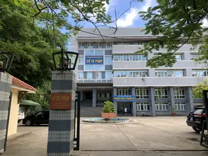 Bức ảnh chụp quang cảnh bên ngoài của một tòa nhà cơ quan chính phủ vào một ngày trời trong. Đây là tòa nhà Sở Tư pháp tỉnh Thanh Hóa, một công trình kiến trúc nhiều tầng, hiện đại với tông màu xám và trắng. Ảnh được chụp từ phía cổng chính nhìn vào.。Các văn bản có thể đọc được trên các biển hiệu bao gồm:。- Trên cột cổng: "SỞ TƯ PHÁP", "34 ĐẠI LỘ LÊ LỢI", "P. ĐIỆN BIÊN - TP THANH HÓA".。- Trên mặt tiền tòa nhà: "ỦY BAN NHÂN DÂN TỈNH THANH HÓA" và "SỞ TƯ PHÁP".。- Phía trên lối vào tầng trệt: "SỞ TƯ PHÁP THANH HÓA", "PHÒNG CÔNG CHỨNG SỐ 1", cùng với địa chỉ "ĐC: 34 ĐẠI LỘ LÊ LỢI - TP. THANH HÓA" và số điện thoại "ĐT: 02373 852 976".。- Biển báo trong sân: "KHU VỰC ĐỂ XE ĐẠP - XE MÁY CỦA KHÁCH" và "KHU VỰC ĐỂ XE Ô TÔ CỦA KHÁCH".。Khung cảnh cho thấy một khoảng sân rộng phía trước tòa nhà, có một đài phun nước nhỏ hình tròn ở giữa. Một vài chiếc xe ô tô đang đỗ trong sân. Một người mặc áo trắng đang đi về phía lối vào Phòng Công chứng. Cây xanh, bao gồm cả cành cây phượng, xuất hiện ở tiền cảnh, tạo nên một không gian xanh mát. Một vài lá cờ Việt Nam được treo trên tòa nhà.。（日本語訳）。晴れた日の政府機関の建物の外観を撮影した写真です。これはタインホア省司法局の建物で、グレーと白を基調とした多層階の近代的な建築物です。写真は正門側から中を向いて撮影されています。看板で読み取れる文言は以下の通りです：。- 門柱：「司法局」、「レロイ大通り34番地」、「ディエンビエン区 - タインホア市」。- 建物の正面：「タインホア省人民委員会」と「司法局」。- 1階入口の上部：「タインホア司法局」、「第1公証役場」、および住所「住所：レロイ大通り34番地 - タインホア市」と電話番号「電話：02373 852 976」。- 庭の看板：「来客用自転車・バイク駐車場」と「来客用自動車駐車場」。風景には、建物の前に広々とした庭があり、中央には小さな円形の噴水があります。庭には数台の自動車が駐車しています。白い服を着た人が一人、公証役場の入口に向かって歩いています。前景にはホウオウボクの枝を含む緑の木々が見え、涼しげな緑の空間を作り出しています。建物には数本のベトナム国旗が掲げられています。