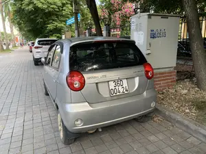 Bối cảnh là vỉa hè của một con phố đô thị, có nhiều cây xanh. Một chiếc ô tô hiệu Chevrolet Spark Lite màu bạc đang đỗ trên vỉa hè lát gạch. Phía sau nó là một hàng dài các xe ô tô khác cũng đang đỗ. Bên cạnh chiếc xe màu bạc là một tủ điện lớn màu xám. Trên tủ điện có ghi các thông tin như: 'TỦ CÔNG TƠ NGOÀI TRỜI', 'TC2.4', 'TRẠ NGỌC TRẠO', và các số điện thoại 'KH.C.B.T 0915.135.119', '1900 6769'. Phía sau tủ điện là hàng rào sắt và giàn hoa giấy màu hồng. Ở phía xa trên vỉa hè, có người đi bộ, trong đó có một người đội nón lá, và có xe máy đang lưu thông trên đường.。（日本語訳）。背景は都市の街路の歩道で、多くの緑の木々があります。一台の銀色のシボレー・スパークライトがタイル張りの歩道に駐車しています。その後ろには、他にも駐車している長い車の列があります。銀色の車の隣には、大きな灰色の配電盤があります。配電盤には、「TỦ CÔNG TƠ NGOÀI TRỜI」（屋外メーターボックス）、「TC2.4」、「TRẠ NGỌC TRẠO」（ゴックチャオ変電所）、そして電話番号「KH.C.B.T 0915.135.119」、「1900 6769」といった情報が書かれています。配電盤の後ろには、鉄の柵とピンクのブーゲンビリアの棚があります。遠くの歩道には歩行者がおり、その中にはノンラーをかぶった人が一人いて、道路ではバイクが通行しています。