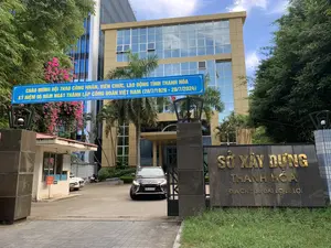 Đây là hình ảnh chụp lối vào trụ sở Sở Xây dựng tỉnh Thanh Hóa, một tòa nhà văn phòng hiện đại, nhiều tầng với mặt tiền bằng kính. Phía trước cổng vào có một tấm biển bằng đá khắc chữ vàng 'SỞ XÂY DỰNG THANH HÓA. ĐỊA CHỈ: 36 ĐẠI LỘ LÊ LỢI'. Phía trên cổng có treo một băng rôn lớn màu xanh với nội dung: 'CHÀO MỪNG HỘI THAO CÔNG NHÂN, VIÊN CHỨC, LAO ĐỘNG TỈNH THANH HÓA KỶ NIỆM 95 NĂM NGÀY THÀNH LẬP CÔNG ĐOÀN VIỆT NAM (28/7/1929 - 28/7/2024)'. Bên trái cổng là một bốt bảo vệ, gần đó có một biển báo nhỏ ghi 'BẢO VỆ'. Trong sân, một chiếc ô tô đang đi qua cổng. Khung cảnh xung quanh có nhiều cây xanh và một giàn hoa giấy màu hồng.。（日本語訳）。これはタインホア省建設局本庁舎の入口を撮影した写真で、ガラス張りのファサードを持つ多層階の近代的なオフィスビルです。入口ゲートの前には、「SỞ XÂY DỰNG THANH HÓA. ĐỊA CHỈ: 36 ĐẠI LỘ LÊ LỢI」（タインホア省建設局。住所：36 レロイ大通り）と金色の文字が刻まれた石の看板があります。ゲートの上には、「CHÀO MỪNG HỘI THAO CÔNG NHÂN, VIÊN CHỨC, LAO ĐỘNG TỈNH THANH HÓA KỶ NIỆM 95 NĂM NGÀY THÀNH LẬP CÔNG ĐOÀN VIỆT NAM (28/7/1929 - 28/7/2024)」（ベトナム労働組合設立95周年（1929年7月28日～2024年7月28日）を記念するタインホア省の労働者、公務員、従業員のためのスポーツ大会を歓迎します）という内容の大きな青い横断幕が掲げられています。ゲートの左側には警備員詰所があり、その近くに「BẢO VỆ」（警備）と書かれた小さな看板があります。敷地内では、一台の車がゲートを通過しています。周囲の景色には多くの緑の木々と、ピンク色のブーゲンビリアの棚があります。