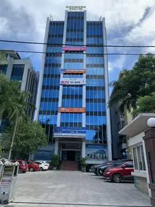 Bức ảnh chụp một tòa nhà văn phòng cao tầng, hiện đại với mặt tiền bằng kính màu xanh dương, nằm ở một khu vực đô thị vào ban ngày. Phía trước tòa nhà là một khoảng sân đỗ xe có nhiều ô tô đang đậu. Bầu trời trong xanh và có mây trắng.。**Chữ viết được ghi lại:**。*   **Trên nóc tòa nhà:** CÔNG TY CP XÂY LẮP DẦU KHÍ THANH HÓA, PVC-TH.。*   **Biển hiệu chính ở lối vào:** TÒA NHÀ DẦU KHÍ, CÔNG TY CỔ PHẦN XÂY LẮP DẦU KHÍ THANH HÓA (PVC-TH), có logo của PETROVIETNAM. Địa chỉ được ghi rõ: TẦNG 10 - TÒA NHÀ DẦU KHÍ - SỐ 38A ĐẠI LỘ LÊ LỢI - P. ĐIỆN BIÊN - TP. THANH HOÁ. Kèm theo là số điện thoại, fax và website.。*   **Các băng rôn trên mặt tiền:**。*   Băng rôn màu hồng: SKY GROUP LOGISTICS, VẬN CHUYỂN CONTAINER NỘI ĐỊA - QUỐC TẾ, 0916.276.888.。*   Băng rôn màu đỏ: CHO THUÊ VĂN PHÒNG, Hotline: 0989.440.888 - 0983.053.858.。*   Băng rôn màu trắng: Global Education, IELTS TEST CENTER, Tầng 6.。*   Băng rôn màu cam: eQ english, IELTS expert, TẦNG 3.。*   **Quảng cáo khác:** Có một tấm áp phích lớn của Vietnam Airlines gần cửa ra vào.。*   **Trên rào chắn tự động (barie):** Barie, Cổng xếp, Hongmen, và các ký tự Trung Quốc (红门开, 开门红).。**Mô tả cảnh quan và hoạt động:**。*   **Loại địa điểm:** Tòa nhà văn phòng thương mại.。*   **Hoạt động cụ thể:** Không có hoạt động rõ ràng của con người ở bên ngoài, nhưng sự hiện diện của các ô tô đang đỗ cho thấy các hoạt động văn phòng và kinh doanh đang diễn ra bên trong tòa nhà.。*   **Các vật thể:** Tòa nhà cao tầng, ô tô, cây cọ, cây xanh, rào chắn tự động, các biển hiệu và băng rôn quảng cáo, đường dây điện.。（日本語訳）。写真は、日中の都市部にある、青いガラスのファサードを持つ高層の近代的なオフィスビルを写しています。ビルの前には多くの車が駐車している駐車場があります。空は青く、白い雲が浮かんでいます。**記録された文字：**。*   **ビルの屋上：** タインホア石油ガス建設株式会社、PVC-TH。*   **入り口のメイン看板：** 石油ガスビル、タインホア石油ガス建設株式会社（PVC-TH）、PETROVIETNAMのロゴあり。住所は明確に記載：タインホア市ディエンビエン坊レロイ大通り38A号 石油ガスビル10階。電話番号、ファックス、ウェブサイトが併記されています。*   **ファサードの横断幕：**。*   ピンク色の横断幕：SKY GROUP LOGISTICS、国内・国際コンテナ輸送、0916.276.888。*   赤色の横断幕：オフィス賃貸、ホットライン：0989.440.888 - 0983.053.858。*   白色の横断幕：Global Education、IELTSテストセンター、6階。*   オレンジ色の横断幕：eQ english、IELTS expert、3階。*   **その他の広告：** 出入り口の近くにベトナム航空の大きなポスターがあります。*   **自動ゲート（遮断機）上：** Barie、Cổng xếp、Hongmen、および中国語の文字（红门开、开门红）。**景観と活動の説明：**。*   **場所の種類：** 商業オフィスビル。*   **具体的な活動：** 屋外では明確な人の活動は見られませんが、駐車している車の存在は、ビル内部でオフィス業務やビジネス活動が行われていることを示しています。*   **物体：** 高層ビル、自動車、ヤシの木、緑の木々、自動ゲート、看板や広告の横断幕、電線。