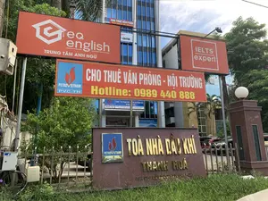 Bức ảnh chụp bên ngoài Toà nhà Dầu khí Thanh Hoá, một tòa nhà văn phòng hiện đại. Phía trước có một tấm biển lớn bằng đá ghi 'TOÀ NHÀ DẦU KHÍ THANH HOÁ' và địa chỉ '38A ĐẠI LỘ LÊ LỢI, THANH HOÁ'. Có rất nhiều biển hiệu quảng cáo cho các dịch vụ khác nhau. Một băng rôn lớn màu đỏ ghi 'CHO THUÊ VĂN PHÒNG - HỘI TRƯỜNG' cùng số điện thoại 'Hotline: 0989 440 888'. Các biển hiệu khác bao gồm 'EQ english TRUNG TÂM ANH NGỮ', 'IELTS expert', và các biển hiệu của 'PETROVIETNAM PVC-TH'. Nhìn qua cửa kính có thể thấy biển hiệu của 'CÔNG TY CỔ PHẦN XÂY LẮP DẦU KHÍ THANH HÓA' và logo của 'Vietnam Airlines'. Khung cảnh cho thấy đây là một khu vực thương mại với cây xanh, hàng rào và một vài chiếc ô tô đang đậu phía sau.。（日本語訳）。タインホア石油ガスビルの外で撮影された写真、近代的なオフィスビルです。正面には「TOÀ NHÀ DẦU KHÍ THANH HOÁ」（タインホア石油ガスビル）と住所「38A ĐẠI LỘ LÊ LỢI, THANH HOÁ」（タインホア、レロイ大通り38A）と書かれた大きな石の看板があります。様々なサービスの広告看板がたくさんあります。大きな赤い横断幕には「CHO THUÊ VĂN PHÒNG - HỘI TRƯỜNG」（オフィス・ホール賃貸）と電話番号「ホットライン：0989 440 888」が書かれています。その他の看板には、「EQ english TRUNG TÂM ANH NGỮ」（EQイングリッシュ英語センター）、「IELTS expert」、そして「PETROVIETNAM PVC-TH」の看板が含まれます。ガラスのドア越しに、「CÔNG TY CỔ PHẦN XÂY LẮP DẦU KHÍ THANH HÓA」（タインホア石油ガス建設株式会社）の看板と「ベトナム航空」のロゴが見えます。この光景は、緑の木々、フェンス、そして後ろに数台の車が駐車している商業地域であることを示しています。