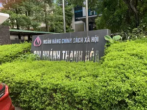 Trên tấm biển hiệu bằng đá có khắc chữ kim loại màu bạc: "NGÂN HÀNG CHÍNH SÁCH XÃ HỘI - CHI NHÁNH THANH HÓA". Bên cạnh có logo của ngân hàng với chữ "VBSP". Đây là khung cảnh bên ngoài trụ sở của Ngân hàng Chính sách xã hội, chi nhánh tỉnh Thanh Hóa. Tấm biển hiệu lớn được đặt giữa một hàng rào cây xanh tươi tốt. Phía sau là tòa nhà của ngân hàng và cây cối. Một phần của chiếc ô tô màu đỏ có thể được nhìn thấy ở góc dưới bên trái.。（日本語訳）。石の看板には「社会政策銀行 - タインホア支店」という銀色の金属文字が刻まれています。その隣には「VBSP」という文字が入った銀行のロゴがあります。これは社会政策銀行タインホア省支店の本社の外の光景です。大きな看板は、青々と茂る生垣の真ん中に設置されています。後ろには銀行の建物と木々があります。左下の隅に赤い自動車の一部が見えます。