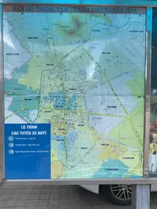 Đây là hình ảnh chụp một tấm bản đồ công cộng được đặt trong khung kim loại và kính. Tiêu đề lớn ở trên cùng ghi 'BẢN ĐỒ TRUNG TÂM THÀNH PHỐ THANH HÓA'. Bản đồ thể hiện chi tiết mạng lưới đường phố, các phường, xã và các địa danh quan trọng trong thành phố Thanh Hóa. Có thể nhận diện các khu vực như Đông Cương, Đông Sơn, Phú Sơn, Tân Sơn, Điện Biên, Ba Đình, Lam Sơn, Đông Vệ, Hàm Rồng, Nam Ngạn, Đông Hương, và huyện Hoằng Hóa. Các địa danh tự nhiên như Núi Hàm Rồng và Sông Mã cũng được thể hiện rõ. Ở góc dưới bên trái của bản đồ là một bảng thông tin màu xanh dương với tiêu đề 'LỘ TRÌNH CÁC TUYẾN XE BUÝT', liệt kê thông tin về các tuyến xe buýt: '01 Ga Thanh Hóa – Cảng Hới', '10 Khu Bình Minh – Ngã 3 Mục sơn', và '16 Ngã 3 Đồng Bởi Như Thanh – Khu DL Hải Tiến'. Tấm bản đồ được đặt ngoài trời trên vỉa hè lát gạch, và một phần bánh xe ô tô có thể được nhìn thấy ở góc dưới bên phải, cho thấy vị trí có thể là một trạm xe buýt hoặc điểm thông tin công cộng ven đường.。（日本語訳）。これは、金属とガラスのフレームに収められた公共の地図を撮影した画像です。上部の大きなタイトルには「BẢN ĐỒ TRUNG TÂM THÀNH PHỐ THANH HÓA」（タインホア市中心部地図）と書かれています。地図は、タインホア市内の道路網、各坊・社、および重要なランドマークを詳細に示しています。ドンクオン、ドンソン、フーソン、タンソン、ディエンビエン、バディン、ラムソン、ドンヴェ、ハムロン、ナムガン、ドンフオン、およびホアンホア県などの地域を識別できます。ハムロン山やマー川などの自然のランドマークもはっきりと示されています。地図の左下には、「LỘ TRÌNH CÁC TUYẾN XE BUÝT」（バス路線図）というタイトルの青い案内板があり、バス路線に関する情報がリストアップされています：「01 タインホア駅 – ホイ港」、「10 ビンミン地区 – ムックソン三叉路」、および「16 ヌータイン・ドンボイ三叉路 – ハイティエン観光区」。地図はタイル張りの歩道上の屋外に設置されており、右下隅には自動車の車輪の一部が見え、その場所がバス停または道端の公共案内所である可能性を示しています。