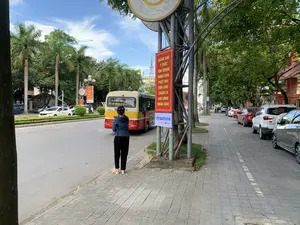 Bản ghi văn bản:。Trên băng rôn tuyên truyền màu đỏ có ghi: 'NÂNG CAO Ý THỨC TÔN TRỌNG NHÂN DÂN, PHÁT HUY DÂN CHỦ, CHĂM LO ĐỜI SỐNG NHÂN DÂN。'.。Trên biển quảng cáo bên dưới có chữ 'vinaphone' và 'VNPT'.。Phía sau xe buýt có ghi lộ trình, một phần có thể đọc được là: 'GA - COOPMART... BIG C - MỚI SẦM SƠN - BÃI TẮM A,B,C,D...'.。Mô tả cảnh quan và hoạt động:。Bối cảnh là một con đường trong thành phố vào một ngày nắng đẹp. Một người phụ nữ đang đứng trên vỉa hè, có vẻ như đang chờ hoặc vẫy một chiếc xe buýt màu vàng và đỏ đang di chuyển trên đường. Dọc theo vỉa hè bên phải là một hàng ô tô đang đỗ. Con đường được bao quanh bởi cây xanh, bao gồm cả những cây cọ cao. Ở phía xa có thể nhìn thấy các tòa nhà, một cần cẩu xây dựng và một tháp ăng-ten. Một cột kim loại ở gần máy ảnh có treo một băng rôn tuyên truyền màu đỏ và một biển quảng cáo của nhà mạng Vinaphone.。（日本語訳）。テキスト記録：。赤いプロパガンダの横断幕には、「人民を尊重する意識を高め、民主主義を発揮し、人民の生活を気遣おう。」と書かれている。下の広告看板には「vinaphone」と「VNPT」の文字がある。バスの後部には路線が書かれており、読み取れる部分には「駅 - コープマート... ビッグC - 新サムソン - A,B,C,Dビーチ...」とある。風景と活動の説明：。背景は晴れた日の都市の道路である。右側の歩道に沿って、駐車している車が一列に並んでいる。道路は緑の木々に囲まれており、その中には背の高いヤシの木も含まれている。