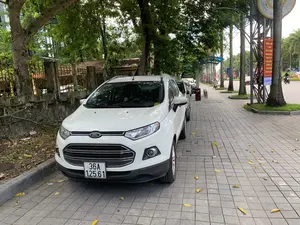 Cảnh quan đô thị với một chiếc ô tô màu trắng hiệu Ford đang đỗ trên vỉa hè lát gạch. Vỉa hè rộng, có hàng cây xanh trồng dọc theo, lá vàng rơi rải rác trên mặt đất. Phía sau chiếc xe là một hàng rào cũ. Ở phía xa trên đường phố, có người đang đi xe máy và các ô tô khác đang lưu thông. Treo trên cột bên đường là một tấm biểu ngữ tuyên truyền màu đỏ có nội dung: 'NÂNG CAO Ý THỨC TÔN TRỌNG NHÂN DÂN, PHÁT HUY DÂN CHỦ, CHĂM LO ĐỜI SỐNG NHÂN DÂN'. Bên dưới có logo 'vinapho'.。（日本語訳）。レンガ敷きの歩道にフォードブランドの白い車が駐車している都市の景観。歩道は広く、沿って街路樹が植えられており、地面には黄色い葉が散らばっている。車の後ろには古いフェンスがある。通りの遠くでは、バイクに乗っている人がおり、他の車が通行している。その下には「vinapho」のロゴがある。