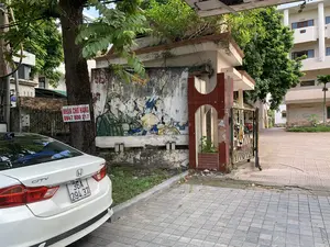 Bối cảnh là lối vào một khu nhà hoặc khu tập thể cũ. Bên trái là một chiếc ô tô màu trắng đang đỗ. Trên một thân cây lớn có treo tấm biển màu đỏ chữ trắng ghi: "NHẬN CHỞ HÀNG 0947 800 217". Bức tường của cổng vào có một bức tranh cổ động đã cũ và phai màu, vẽ hình một người lính đội mũ cối và những người dân. Cổng vào có trụ ốp gạch men màu đỏ và một cánh cổng sắt. Sân được lát gạch. Xung quanh có nhiều cây xanh và phía sau là một tòa nhà nhiều tầng.。（日本語訳）。背景は、ある住宅地または古い集合住宅の入り口です。左側には白い車が一台駐車しています。大きな木の幹に、「貨物輸送承ります 0947 800 217」と白字で書かれた赤い看板が掛けられています。入り口の壁には、古びて色褪せたプロパガンダの絵があり、ヘルメットをかぶった兵士と人々が描かれています。入り口には、赤いタイル張りの柱と鉄の門があります。中庭はタイル張りです。周囲には多くの緑の木々があり、後ろには多層階の建物があります。