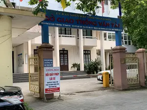 Bức ảnh chụp lối vào của một tòa nhà văn phòng chính phủ. Đây là trụ sở của Sở Giao thông Vận tải tỉnh Thanh Hóa. Phía trên cổng có một biển hiệu lớn màu xanh với dòng chữ đỏ "SỞ GIAO THÔNG VẬN TẢI" và dòng chữ trắng "ỦY BAN NHÂN DÂN TỈNH THANH HÓA". Biển hiệu cũng cung cấp thông tin chi tiết: "Đ/C: 42 Đại Lộ Lê Lợi - P. Điện Biên - TP. Thanh Hóa * ĐT: 0373.852 360 - Fax: 0373. 855 129 * Website: sgtvt.thanhhoa.gov.vn - Emai: sgtvt.@thanhhoa.gov.vn".。Tại cổng, có một biển báo nhỏ màu xanh yêu cầu: "YÊU CẦU QUÝ KHÁCH XUỐNG XE XUẤT TRÌNH GIẤY TỜ". Bên cạnh đó là một biển "THÔNG BÁO" lớn hơn, thông báo rằng dịch vụ "ĐỔI GIẤY PHÉP LÁI XE" đã được "CHUYỂN VỀ TRUNG TÂM PHỤC VỤ HÀNH CHÍNH CÔNG TỈNH THANH HÓA" tại địa chỉ mới: "Số: 28 Đại Lộ Lê Lợi, P. Điện Biên, TP. Thanh Hóa".。Khung cảnh cho thấy một tòa nhà nhiều tầng, một cổng sắt màu vàng đang mở, một thanh chắn barie tự động, và một phần của hai chiếc ô tô đang đỗ. Xung quanh có nhiều cây xanh. Không có hoạt động nào của con người được ghi lại, chỉ có khung cảnh tĩnh của lối vào một cơ quan hành chính.。（日本語訳）。政府庁舎の入り口を撮影した写真です。これはタインホア省交通運輸局の本庁舎です。門の上には、赤い文字で「SỞ GIAO THÔNG VẬN TẢI」（交通運輸局）、白い文字で「ỦY BAN NHÂN DÂN TỈNH THANH HÓA」（タインホア省人民委員会）と書かれた大きな青い看板があります。看板には詳細情報も記載されています。「住所：タインホア市ディエンビエン区レロイ大通り42番地 * 電話：0373.852 360 - ファックス：0373. 855 129 * ウェブサイト：sgtvt.thanhhoa.gov.vn - Eメール：sgtvt.@thanhhoa.gov.vn」。門には、「YÊU CẦU QUÝ KHÁCH XUỐNG XE XUẤT TRÌNH GIẤY TỜ」（お客様は降車し、身分証明書を提示してください）と要求する小さな青い看板があります。その隣には、より大きな「THÔNG BÁO」（お知らせ）の看板があり、「ĐỔI GIẤY PHÉP LÁI XE」（運転免許証の交換）サービスが、新しい住所「タインホア市ディエンビエン区レロイ大通り28番地」にある「TRUNG TÂM PHỤC VỤ HÀNH CHÍNH CÔNG TỈNH THANH HÓA」（タインホア省公共行政サービスセンター）に移転したことを知らせています。周りには多くの緑の木々があります。人の活動は記録されておらず、行政機関の入り口の静かな風景だけがあります。