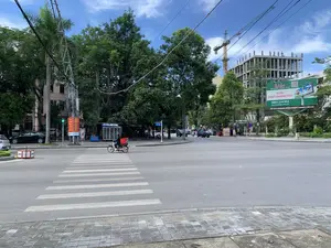 Bức ảnh chụp một ngã tư đường phố vào một ngày nắng. Khung cảnh cho thấy một con đường nhựa rộng rãi với vạch kẻ đường dành cho người đi bộ. Một người đang lái xe máy chở một chiếc sọt nhựa màu đỏ ở phía sau, đi qua ngã tư. Dọc hai bên đường là hàng cây xanh và các tòa nhà. Ở phía xa bên phải, một tòa nhà cao tầng đang được xây dựng, với một cần cẩu lớn màu vàng và xanh lá cây hoạt động bên cạnh. Có một biển quảng cáo lớn màu xanh lá cây với nội dung: 'Hatuba Advertising - Media - Event', 'VỊ TRÍ CHÀO QUẢNG CÁO', số điện thoại '0903 214 932' và trang web 'www.hatuba.com.vn'. Một biển báo tên đường màu xanh dương ghi 'ĐẠI LỘ LÊ LỢI'. Ngoài ra còn có một cột đèn tín hiệu giao thông đang hiển thị đèn xanh và một biển báo màu đỏ có logo 'VNPT Vinaphone' nhưng nội dung chi tiết không đọc được rõ.。（日本語訳）。晴れた日の街の交差点を撮影した写真です。一人の人が、後ろに赤いプラスチック製のカゴを乗せたバイクを運転し、交差点を渡っています。道路の両側には緑の木々と建物が並んでいます。右奥では高層ビルが建設中で、その隣では黄色と緑色の大きなクレーンが稼働しています。「Hatuba Advertising - Media - Event」、「VỊ TRÍ CHÀO QUẢNG CÁO（広告募集中の場所）」、電話番号「0903 214 932」、ウェブサイト「www.hatuba.com.vn」という内容の大きな緑色の広告看板があります。「ĐẠI LỘ LÊ LỢI（レ・ロイ大通り）」と書かれた青い道路標識があります。その他にも、青信号を表示している信号機と、「VNPT Vinaphone」のロゴが入った赤い看板がありますが、詳細な内容ははっきりと読み取れません。