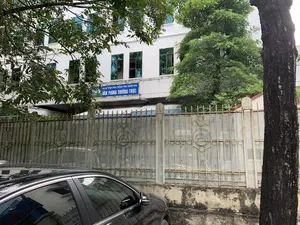 Bản ghi văn bản từ biển hiệu màu xanh trên tòa nhà: 'BAN AN TOÀN GIAO THÔNG TỈNH THANH HÓA - VĂN PHÒNG THƯỜNG TRỰC'.。Bối cảnh là phía trước một tòa nhà văn phòng, cụ thể là trụ sở Văn phòng Thường trực của Ban An toàn Giao thông tỉnh Thanh Hóa. Tòa nhà có màu trắng, nhiều tầng với các cửa sổ kính màu xanh. Phía trước tòa nhà được bao quanh bởi một hàng rào kim loại. Ở tiền cảnh, có một chiếc ô tô màu đen đang đỗ và nhiều cây xanh. Một thân cây lớn nằm ở phía bên phải của bức ảnh. Không có hoạt động cụ thể nào của con người được nhìn thấy.。（日本語訳）。建物の青い看板からのテキストの書き起こし：「タインホア省交通安全委員会 - 常設事務局」。背景はオフィスビルの前で、具体的にはタインホア省交通安全委員会の常設事務局の本部です。建物は白色で、青いガラス窓のある多層階建てです。建物の前は金属製のフェンスで囲まれています。前景には、駐車している黒い車と多くの緑の木々があります。写真の右側には大きな木の幹があります。特定の人の活動は見られません。