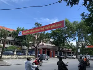 Bức ảnh chụp một con đường tại thành phố Thanh Hóa vào ban ngày, dưới bầu trời trong xanh. Một băng rôn lớn màu đỏ được treo ngang qua đường với dòng chữ màu vàng: 'CHĂM SÓC, PHỤNG DƯỠNG NGƯỜI CAO TUỔI LÀ TRUYỀN THỐNG TỐT ĐẸP CỦA DÂN TỘC。'. Phía sau là một tòa nhà lớn, có thể là một cơ quan hành chính nhà nước, với biển hiệu có các dòng chữ như 'ỦY BAN NHÂN DÂN TỈNH THANH H...', '...ĐẦU TƯ XÂY DỰNG CÁC CÔNG TRÌNH...' và '...PHƯỜNG TÂN SƠN - TP. THANH HÓA'. Trên đường phố, nhiều người đang điều khiển xe máy tham gia giao thông. Một số người mặc áo chống nắng và đội mũ. Nhiều xe ô tô đang đỗ dọc bên đường. Cây xanh được trồng dọc hai bên đường.。（日本語訳）。青空の下、日中のタインホア市のある道を写した写真。道を横切って大きな赤い横断幕が掲げられており、黄色い文字で「高齢者の世話をし、養うことは民族の良き伝統です。」と書かれている。後ろには大きな建物があり、おそらく国の行政機関で、「タインホア省人民委員会…」、「…各工事の建設投資…」、「…タンソン坊 - タインホア市」といった文言が書かれた看板がある。路上では、多くの人々がバイクを運転して交通に参加している。何人かは日焼け防止の上着を着て、帽子をかぶっている。多くの自動車が道端に沿って駐車している。道の両脇には緑の木々が植えられている。