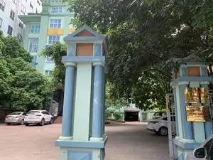 Bức ảnh chụp lối vào của một tòa nhà văn phòng. Ở phía trước là hai cột cổng lớn, được sơn màu xanh lam và xanh lá nhạt. Bên trong cổng là một sân rộng lát gạch, có một vài chiếc ô tô đang đỗ. Phía sau là một tòa nhà nhiều tầng màu xanh lá cây, được bao quanh bởi cây cối um tùm. Trên cột cổng bên phải có gắn các tấm biển hiệu bằng kim loại màu vàng. Nội dung trên một tấm biển có thể đọc được là: "TỔNG CÔNG TY CỔ PHẦN BẢO HIỂM BƯU ĐIỆN. CÔNG TY BẢO HIỂM BƯU ĐIỆN THANH HÓA. MST: 0100774631-014. Đ/C: TẦNG 5, SỐ 11 HẠC THÀNH, P. ĐIỆN BIÊN, TP. THANH HÓA. ĐIỆN THOẠI: 02373 714 234". Trên tường của tòa nhà phía sau cũng có một biển hiệu màu vàng với logo và dòng chữ "ISO 9001:2008". Khung cảnh cho thấy đây là một khu vực văn phòng vào một ngày bình thường, không có hoạt động rõ rệt của con người.。（日本語訳）。オフィスビルの入り口を写した写真です。正面には、青と薄緑色に塗られた2本の大きな門柱があります。門の内側にはタイル敷きの広い中庭があり、数台の車が駐車しています。後方には緑色の多層階建ての建物があり、緑豊かな木々に囲まれています。右側の門柱には、黄色の金属製の看板がいくつか取り付けられています。読み取れる看板の一つには、「郵便保険株式会社総公社。タインホア郵便保険会社。税コード：0100774631-014。住所：タインホア市ディエンビエン区ハクタン通り11番地5階。電話番号：02373 714 234」と書かれています。後方の建物の壁にも、ロゴと「ISO 9001:2008」という文字が入った黄色の看板があります。この光景は、ここがオフィス街のありふれた一日であり、目立った人の活動がないことを示しています。