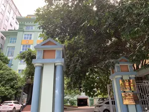 Bức ảnh chụp lối vào của một tòa nhà văn phòng. Phía trước là cổng vào với hai cột trụ lớn sơn màu xanh lam và trắng. Phía sau cổng là một tòa nhà nhiều tầng sơn màu xanh lục, có nhiều cửa sổ. Một cây lớn với tán lá xum xuê che khuất một phần tòa nhà. Trong sân có một vài chiếc ô tô đang đỗ. Không có hoạt động nào của con người đang diễn ra. Trên tòa nhà và cột cổng có các biển hiệu với nội dung như sau: Trên nóc tòa nhà có chữ 'TÒA NHÀ'. Trên tường tòa nhà có biển hiệu màu vàng với logo và dòng chữ 'ISO 9001:2008'. Trên cột cổng bên phải có gắn một tấm biển kim loại màu vàng ghi rõ: 'CÔNG TY BẢO HIỂM BƯU ĐIỆN THANH HÓA', 'MST: 0100774631-01', 'Đ/C: TẦNG 5, SỐ 11 HẠC THÀNH, P. ĐIỆN BIÊN, TP. THANH HÓA', 'ĐIỆN THOẠI: 02373 718 234'.。（日本語訳）。オフィスビルの入り口の写真です。前方には、青と白に塗られた2本の大きな柱がある門があります。門の後ろには、緑色に塗られた多層階の建物があり、多くの窓があります。葉が茂った大きな木が建物の一部を覆い隠しています。敷地内には数台の車が駐車しています。人の活動は見られません。建物と門の柱には、次のような内容の看板があります。建物の屋上には「TÒA NHÀ」という文字があります。建物の壁には、ロゴと「ISO 9001:2008」という文字が入った黄色い看板があります。右側の門の柱には、次のように明記された黄色い金属製のプレートが取り付けられています。「タインホア郵便保険会社」、「税コード: 0100774631-01」、「住所: タインホア市ディエンビエン坊ハク・タイン通り11番地5階」、「電話: 02373 718 234」。