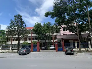 Bản ghi văn bản:。- Trên mái tòa nhà có một tấm biển lớn màu đỏ ghi chữ: "ỦY BAN NHÂN DÂN... BAN QUẢN LÝ DỰ ÁN ĐẦU TƯ XÂY DỰNG CÁC... HẠC THÀNH - PHƯỜNG TÂN SƠN". Một phần của dòng chữ bị cây che khuất.。- Trên cột cổng bên phải có một biển số nhà màu xanh ghi "06 HẠC THÀNH".。- Ở phía bên phải của lối vào, có một tấm biển màu đỏ khác với trích dẫn: ""TỈNH THANH HÓA PHẢI TRỞ NÊN MỘT TỈNH KIỂU..."", đây là một câu nói của Chủ tịch Hồ Chí Minh.。Mô tả cảnh quan và hoạt động:。- Loại địa điểm: Đây là một tòa nhà văn phòng, là trụ sở của một cơ quan nhà nước, tọa lạc tại số 06 đường Hạc Thành, thành phố Thanh Hóa. Dựa trên tấm biển, đây là trụ sở của Ban Quản lý dự án đầu tư xây dựng.。- Hoạt động: Khung cảnh tĩnh lặng, không có hoạt động rõ rệt của con người. Có nhiều ô tô đang đỗ trong sân của tòa nhà, cho thấy đây có thể là một ngày làm việc.。- Các vật thể trong ảnh: Một tòa nhà lớn nhiều tầng với nhiều cửa sổ kính. Phía trước là một khoảng sân lát gạch được sử dụng làm bãi đỗ xe. Có một cổng sắt màu xanh lam và hàng rào bao quanh. Nhiều cây xanh lớn được trồng trong khuôn viên. Bầu trời trong xanh với vài gợn mây.。（日本語訳）。テキスト記録：。- 建物の屋根の上には、「人民委員会... 各建設投資プロジェクト管理委員会... ハク・タイン - タン・ソン区」と書かれた大きな赤い看板があります。文字の一部は木で隠されています。- 右側の門柱には、「06 ハク・タイン」と書かれた青い番地プレートがあります。- 入り口の右側には、「『タインホア省は模範的な省にならなければならない...』」という引用文が書かれた別の赤い看板があります。これはホー・チ・ミン主席の言葉です。風景と活動の説明：。- 場所の種類：これはオフィスビルであり、国家機関の本部で、タインホア市ハク・タイン通り06番地に位置しています。看板によると、ここは建設投資プロジェクト管理委員会の本部です。- 活動：静かな光景で、人の明確な活動はありません。建物の敷地内には多くの車が駐車しており、これが就業日であることを示している可能性があります。- 写真の中の物体：多くのガラス窓がある大きな多層階の建物。前面には駐車場として使用されているタイル敷きの中庭があります。青い鉄の門と周囲を囲むフェンスがあります。敷地内には多くの大きな緑の木が植えられています。空は青く澄んでおり、いくつかの筋雲があります。