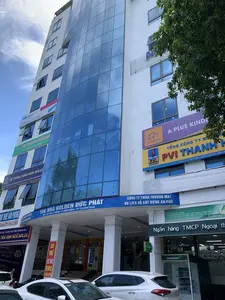 Bức ảnh chụp một tòa nhà văn phòng cao tầng, hiện đại có tên là 'TÒA NHÀ GOLDEN ĐỨC PHÁT' tại địa chỉ '09 HẠC THÀNH - P ĐIỆN BIÊN - TP THANH HÓA'. Tòa nhà có mặt tiền sơn trắng và một phần lớn được ốp kính màu xanh lam. Đây là một tòa nhà thương mại, là nơi đặt trụ sở của nhiều công ty và tổ chức khác nhau, có thể nhận thấy qua các biển hiệu: 'Ngân hàng TMCP Ngoại thương Việt Nam (Vietcombank)' có một cây ATM 24h ở tầng trệt và một biểu ngữ 'CHÀO MỪNG LỄ KỶ NIỆM 60 NĂM THÀNH LẬP...'; 'TỔNG CÔNG TY BẢO HIỂM PVI THANH HÓA'; 'HỆ THỐNG MẦM NON APLUS' và 'TRUNG TÂM ANH NGỮ APLUS'; 'VĂN PHÒNG ĐẠI LÝ KHU VỰC THANH HÓA' của MB Ageas; 'CÔNG TY TNHH THƯƠNG MẠI DU LỊCH VÀ XÂY DỰNG AN PHÚ'. Ngoài ra còn có các biển hiệu 'CHO THUÊ VĂN PHÒNG'. Phía trước tòa nhà, trên đường phố, có một vài chiếc ô tô đang đỗ. Khung cảnh cho thấy một con phố sầm uất với các hoạt động kinh doanh thường ngày.。（日本語訳）。写真は、住所「タインホア市ディエンビエン区ハクタン通り09番地」にある「GOLDEN DUC PHATビル」という名の、高層で近代的なオフィスビルを写しています。ビルのファサードは白く塗装されており、大部分が青いガラスで覆われています。これは商業ビルであり、様々な企業や組織の本拠地となっており、それは以下の看板から見て取れます：「ベトナム外商銀行（ベトコムバンク）」は、1階に24時間対応のATMがあり、「設立60周年記念式典を歓迎します...」という横断幕を掲げています。「PVI保険総公社タインホア」。「APLUS幼稚園システム」と「APLUS英語センター」。MB Ageasの「タインホア地域代理店事務所」。「AN PHU貿易観光建設有限会社」。その他に、「貸事務所」という看板もあります。ビルの前の路上には、数台の車が駐車しています。この光景は、日常的な商業活動が行われている賑やかな通りを示しています。