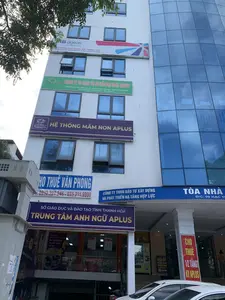 Bức ảnh chụp mặt tiền của một tòa nhà văn phòng cao tầng hiện đại tại thành phố Thanh Hóa. Tòa nhà có kiến trúc kết hợp giữa tường sơn trắng và mặt dựng kính màu xanh. Dựa trên các biển hiệu, địa chỉ của tòa nhà là số 09 đường Hạc Thành, phường Điện Biên, thành phố Thanh Hóa.。Nhiều biển hiệu của các công ty và tổ chức được treo trên tòa nhà, bao gồm:。- 'MB ageas BẢO HIỂM NHÂN THỌ - VĂN PHÒNG ĐẠI LÝ KHU VỰC THANH HÓA' (Tầng 4).。- 'CÔNG TY CP DỊCH VỤ TƯ VẤN ĐẠI NAM GROUP'.。- 'HỆ THỐNG MẦM NON APLUS'.。- 'CÔNG TY TNHH ĐẦU TƯ XÂY DỰNG VÀ PHÁT TRIỂN HẠ TẦNG HỢP LỰC'.。- 'SỞ GIÁO DỤC VÀ ĐÀO TẠO TỈNH THANH HÓA - TRUNG TÂM ANH NGỮ APLUS'.。Ngoài ra còn có các băng rôn cho thuê mặt bằng như 'CHO THUÊ VĂN PHÒNG' với số điện thoại '0932.327.246 - 035.211.9999' và 'CHO THUÊ 1/2 TẦNG 1 KV APLUS'.。Ở tầng trệt có lối vào của Trung tâm Anh ngữ Aplus, một quán cà phê có biển 'Café 09 HẠC THÀNH CÓ MÁY LẠNH SẢNH TẦNG 1', và một máy bán hàng tự động. Phía trước tòa nhà có cây xanh và một vài chiếc ô tô đang đỗ. Khung cảnh cho thấy đây là một tòa nhà văn phòng, nơi diễn ra các hoạt động kinh doanh, giáo dục và dịch vụ.。（日本語訳）。この写真はタインホア市にある近代的な高層オフィスビルの正面を撮影したものです。このビルは、白い塗装の壁と青いガラスのカーテンウォールを組み合わせた建築様式です。看板によると、ビルの住所はタインホア市ディエンビエン区ハク・タイン通り9番地です。ビルには、以下を含む多くの企業や組織の看板が掲げられています。- 「MBアジェアス生命保険 - タインホア地域代理店事務所」（4階）。- 「ダイナムグループコンサルティングサービス株式会社」。- 「APLUS幼児教育システム」。- 「ホップルック建設投資及びインフラ開発有限会社」。- 「タインホア省教育訓練局 - APLUS英語センター」。その他にも、「オフィス貸出」電話番号「0932.327.246 - 035.211.9999」や「APLUSエリア1階の半分を賃貸」といったテナント募集の横断幕があります。1階にはAPLUS英語センターの入り口、「カフェ09ハク・タイン 1階ロビー冷房完備」という看板のあるカフェ、そして自動販売機があります。ビルの前には緑の木々があり、数台の車が駐車しています。この光景は、ここがビジネス、教育、サービスの活動が行われるオフィスビルであることを示しています。