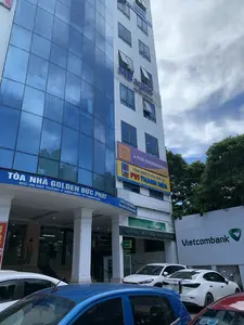Bức ảnh chụp một tòa nhà văn phòng cao tầng, hiện đại vào ban ngày tại một khu vực đô thị. Đây là "TÒA NHÀ GOLDEN ĐỨC PHÁT", có địa chỉ tại "Đ/C: 09 HẠC THÀNH - P ĐIỆN BIÊN - TP THANH HÓA". Tòa nhà có mặt tiền được ốp kính màu xanh lam và tường sơn trắng.。Trên tòa nhà có nhiều biển hiệu của các doanh nghiệp khác nhau:。- Tầng trệt là chi nhánh của "Ngân hàng TMCP Ngoại thương Việt Nam" (Vietcombank). Có một biển hiệu nhỏ chỉ dẫn đến cây ATM và một băng rôn với nội dung "CHÀO MỪNG LỄ KỶ NIỆM 60 NĂM THÀNH LẬP NGÂN HÀNG... VÀ ĐÓN NHẬN DANH HIỆU ANH HÙNG LAO ĐỘNG".。- Các tầng trên có biển hiệu của các công ty: "MB ageas", "Fubon Life", "Vietrantour", trường mầm non "A PLUS KINDERGARTEN", "TỔNG CÔNG TY BẢO HIỂM PVI - PVI THANH HÓA" (địa chỉ tại Tầng 2), và "CÔNG TY TNHH THƯƠNG MẠI DU LỊCH VÀ XÂY DỰNG AN PHÚ".。Hoạt động chính là cảnh sinh hoạt đô thị, với nhiều xe ô tô đang đỗ ở phía trước tòa nhà, cho thấy đây là nơi làm việc và giao dịch. Một người phụ nữ đang đứng gần lối vào của ngân hàng. Phía sau và bên phải tòa nhà có cây xanh. Bầu trời có mây.。（日本語訳）。この写真は、ある都市部で日中に撮影された、高層で近代的なオフィスビルを写したものです。これは「GOLDEN DUC PHATビル」で、住所は「タインホア市ディエンビエン区ハクタン通り09番地」です。ビルの正面は青いガラスで覆われ、壁は白く塗られています。ビルには、様々な企業の看板が多数掲げられています：。- 1階は「ベトナム外商銀行」（Vietcombank）の支店です。ATMへの案内を示す小さな看板と、「銀行設立60周年記念式典...及び労働英雄の称号授与を歓迎します」という内容の横断幕があります。- 上の階には、「MB ageas」、「Fubon Life」、「Vietrantour」、幼稚園「A PLUS KINDERGARTEN」、「PVI保険総公社 - PVIタインホア」（住所：2階）、そして「アンフー建設・旅行・貿易有限会社」といった企業の看板があります。主な活動は都市の生活風景であり、ビルの前に多くの自動車が駐車していることから、ここが職場であり取引の場であることがわかります。一人の女性が銀行の入り口近くに立っています。ビルの後ろと右側には緑の木々があります。空には雲があります。