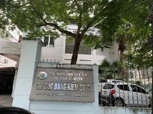 Bức ảnh chụp lối vào của một tòa nhà văn phòng. Ở phía trước có một tấm biển lớn bằng đá, trên đó có khắc chữ kim loại màu bạc. Nội dung trên biển hiệu được ghi lại như sau:。'CỤC ĐĂNG KIỂM VIỆT NAM。VIETNAM REGISTER。CHI CỤC ĐĂNG KIỂM SỐ 12。ĐC: 07 HẠC THÀNH - TP THANH HOA'。Đây là trụ sở của Chi cục Đăng kiểm số 12, một cơ quan nhà nước, tọa lạc tại thành phố Thanh Hóa. Phía sau tấm biển là một tòa nhà màu trắng nhiều tầng. Một cây lớn với tán lá xanh tươi che khuất một phần mặt tiền của tòa nhà. Khuôn viên được bao quanh bởi hàng rào sắt. Bên trong sân, có một chiếc ô tô màu trắng đang đỗ. Không có hoạt động cụ thể nào của con người diễn ra, chỉ là một khung cảnh tĩnh tại một cơ quan hành chính.。（日本語訳）。オフィスビルの入り口を撮影した写真です。手前には大きな石の看板があり、その上には銀色の金属文字が刻まれています。看板の内容は次のように記されています。「ベトナム登録検査局。VIETNAM REGISTER。第12号登録検査支局。住所：タインホア市ハクタン通り07番地」。これはタインホア市に所在する国家機関である第12号登録検査支局の本部です。看板の後ろには、白い多層階の建物があります。青々とした葉を茂らせた大きな木が、建物の正面の一部を覆っています。敷地は鉄柵で囲まれています。庭の内側には、白い自動車が1台駐車しています。特定の人の活動は見られず、行政機関の静かな光景が広がっているだけです。