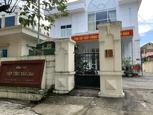 Hình ảnh chụp lối vào của một tòa nhà công vụ, là Trụ sở Tiếp công dân tỉnh Thanh Hóa. Tòa nhà chính là một công trình nhiều tầng, sơn màu trắng, có cổng sắt màu đen. Các dòng chữ trên các biển hiệu có thể được ghi lại như sau:。- Biển hiệu lớn màu nâu bên trái cổng: "TRỤ SỞ TIẾP CÔNG DÂN TỈNH", "ĐỊA CHỈ: SỐ 05 HẠC THÀNH - TP. THANH HÓA".。- Băng rôn màu đỏ phía trên cổng: "TRỤ SỞ TIẾP CÔNG DÂN ... H HÓA".。- Biển số nhà màu vàng trên cột cổng: "05 HẠC THÀNH".。- Biển báo nhỏ màu đỏ bên trong sân: "CÔNG DÂN KHÔNG ĐỖ XE TRONG TRỤ SỞ".。Bên trong sân sau cánh cổng có thể thấy một vài chiếc xe máy đang đỗ và nhiều chậu cây cảnh. Khung cảnh yên tĩnh, không có hoạt động nào rõ rệt của con người đang diễn ra.。（日本語訳）。タインホア省人民受付本庁舎という、ある役所の建物の入り口を撮影した写真です。本館は多層階建ての建物で、白く塗られており、黒い鉄の門があります。看板の文字は次のように記録できます。- 門の左側にある大きな茶色の看板：「省人民受付本庁舎」、「住所：タインホア市ハクタン通り05番地」。- 門の上にある赤い横断幕：「人民受付本庁舎...ホア省」。- 門柱にある黄色の番地表示：「ハクタン05」。- 敷地内の小さな赤い看板：「市民は庁舎内に駐車しないでください」。門の奥の敷地内には、数台のバイクが駐車しており、多くの鉢植えの植物が見えます。静かな光景で、目立った人の活動は見られません。