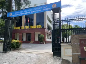 Bản ghi chữ viết: Trên băng rôn lớn màu xanh dương treo ngang cổng vào có dòng chữ: 'NHIỆT LIỆT CHÀO MỪNG KỶ NIỆM 95 NĂM NGÀY THÀNH LẬP CÔNG ĐOÀN VIỆT NAM (28/7/1929 - 28/7/2024)。'. Trên tòa nhà có dòng chữ nổi màu vàng 'THANH TRA TỈNH THANH HÓA'.。Mô tả cảnh vật và hoạt động: Hình ảnh chụp lối vào trụ sở của một cơ quan nhà nước, cụ thể là Thanh tra tỉnh Thanh Hóa. Đây là một tòa nhà văn phòng nhiều tầng, hiện đại. Cổng sắt lớn màu đen đang mở, dẫn vào một khoảng sân rộng phía trước tòa nhà. Hai lá cờ Tổ quốc Việt Nam được treo ở mặt tiền. Một băng rôn lớn được treo để chào mừng một sự kiện kỷ niệm. Xung quanh có cây cọ và các bụi cây hoa trang trí. Khung cảnh yên tĩnh, không có người nào được nhìn thấy trong ảnh.。（日本語訳）。文字の記録：入口の門に横に掲げられた大きな青い横断幕には、「ベトナム労働組合設立95周年記念（1929/7/28 - 2024/7/28）を熱烈に歓迎します。」という文言がある。建物には「タインホア省監察局」という黄色の浮き文字がある。風景と活動の説明：画像は、ある国家機関、具体的にはタインホア省監察局の本庁舎の入口を写したものである。これは現代的な多層階のオフィスビルである。大きな黒い鉄の門が開いており、建物の前にある広い中庭に通じている。周囲にはヤシの木や装飾用の花の茂みがある。風景は静かで、写真には誰も写っていない。