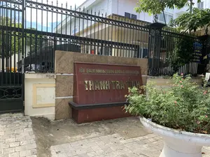 Bức ảnh chụp phía trước một tòa nhà công vụ, có thể là một cơ quan nhà nước. Nổi bật ở trung tâm là một tấm biển lớn bằng đá màu nâu đỏ, trên đó có khắc chữ mạ vàng. Dòng chữ trên ghi 'ỦY BAN NHÂN DÂN TỈNH THANH HÓA', và dòng chữ lớn hơn bên dưới là 'THANH TRA TỈNH'. Tấm biển này được đặt trên vỉa hè lát gạch. Phía sau tấm biển là một hàng rào sắt cao màu đen, kiên cố, ngăn cách với khuôn viên bên trong. Bên trong hàng rào có thể thấy một tòa nhà nhiều tầng màu trắng và cây xanh. Bên phải tấm biển có một chậu cây cảnh lớn màu trắng, trồng một bụi hoa hồng đang nở hoa màu hồng. Khung cảnh yên tĩnh, không có người.。（日本語訳）。写真は、ある公務用の建物の前で撮影されたもので、おそらくは国家機関でしょう。中央に目立つのは、赤褐色の石でできた大きな看板で、その上には金メッキの文字が刻まれています。上の行には「ỦY BAN NHÂN DÂN TỈNH THANH HÓA」、その下の大きな文字の行には「THANH TRA TỈNH」と書かれています。この看板はタイル張りの歩道の上に置かれています。看板の後ろには、頑丈な黒い高い鉄柵があり、内側の敷地とを隔てています。柵の内側には、白い多層階の建物と緑の木々が見えます。看板の右側には、大きな白い植木鉢があり、ピンク色の花を咲かせているバラの茂みが植えられています。辺りは静かで、人はいません。