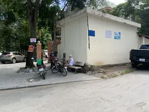 Bối cảnh là một góc phố tại Việt Nam, có vẻ là ở khu dân cư. Trên vỉa hè, một người thợ đeo khẩu trang đang cắt tóc cho một khách hàng, tạo thành một tiệm cắt tóc ngoài trời đơn giản với ghế, quạt điện và gương. Một người đàn ông lớn tuổi khác mặc áo may ô trắng đang ngồi trên ghế đá gần đó. Xung quanh có cây xanh, một chiếc ô tô màu bạc, một xe bán tải màu đen, một xe máy và một xe đạp điện đang đỗ. Các thông tin bằng chữ viết có thể nhận dạng bao gồm: Trên thân cây có biển báo "BÁN NHÀ 28 PHẠM VĂN HINH" và một biển khác ghi "CHO THUÊ NHÀ" với số điện thoại "0907 200 183". Trên tường của một tòa nhà có dán các biển hiệu: một biển của "CÔNG TY CP DP Y TẾ THANH HÓA, NHÀ THUỐC THĂNG HOA, SỐ 1 PHAN VĂN HINH P. ĐIỆN BIÊN TP. THANH HÓA, SỐ GP: 209/NT/TH-GPP/KKD...". Một biển khác màu xanh ghi "PSA RENT STATION". Trên chiếc ghế đá có dòng chữ "SUBET THẾ GIỚI CASINO TRONG TAY BẠN". Một lá cờ màu cam được cắm trên cột gạch.。（日本語訳）。背景はベトナムのある街角で、住宅街のようです。歩道では、マスクをした理容師が客の髪を切っており、椅子、扇風機、鏡を備えた簡単な屋外理髪店を形成しています。近くの石のベンチには、白いタンクトップを着た別の年配の男性が座っています。周りには緑の木々、銀色の乗用車、黒いピックアップトラック、バイク、電動自転車が駐車しています。識別可能な文字情報には以下が含まれます：木の幹には「BÁN NHÀ 28 PHẠM VĂN HINH（ファム・ヴァン・ヒン通り28番地、家売ります）」という看板と、電話番号「0907 200 183」が記載された「CHO THUÊ NHÀ（家貸します）」という別の看板があります。ある建物の壁にはいくつかの看板が貼られています。一つは「CÔNG TY CP DP Y TẾ THANH HÓA, NHÀ THUỐC THĂNG HOA, SỐ 1 PHAN VĂN HINH P. ĐIỆN BIÊN TP. THANH HÓA, SỐ GP: 209/NT/TH-GPP/KKD...（タインホア医療製薬株式会社、タンホア薬局、タインホア市ディエンビエン区ファン・ヴァン・ヒン通り1番地、営業許可番号：209/NT/TH-GPP/KKD...）」の看板です。もう一つの青い看板には「PSA RENT STATION」と書かれています。石のベンチには「SUBET THẾ GIỚI CASINO TRONG TAY BẠN（SUBET あなたの手の中のカジノの世界）」という文字があります。レンガの柱にはオレンジ色の旗が立てられています。