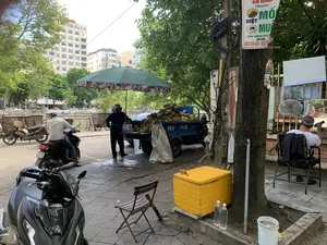 Bối cảnh là một vỉa hè trên một con phố tại một khu đô thị. Một người phụ nữ đang đứng bán dừa bên cạnh một chiếc xe ba gác màu xanh chở đầy dừa tươi. Phía trên có một chiếc ô lớn màu xanh lá cây để che nắng. Gần đó, một người đàn ông mặc áo phông trắng đang ngồi trên ghế và nhìn vào điện thoại di động, bên cạnh có một chiếc quạt và gương, trông giống một tiệm cắt tóc vỉa hè. Một người đàn ông khác đội mũ bảo hiểm đang ngồi trên xe máy. Trên vỉa hè còn có các vật dụng như một chiếc ghế xếp, một chiếc điếu cày, một thùng gỗ sơn màu vàng và vài chai nước. Phía sau là những tòa nhà cao tầng và nhiều cây xanh. Một cột điện có dây điện chằng chịt và treo biển quảng cáo.。Nội dung văn bản trên biển quảng cáo có thể đọc được là: 'AN BÌNH - Chuyên: DIỆT MỐI, DIỆT MUỖI, BẢO HÀNH DÀI HẠN, HOTLINE: 0976 754 555 - 0237...'. Trên thùng xe ba gác có chữ 'NEW H...E'. Ở phía trước có một chiếc xe máy hiệu 'HONDA' đang đỗ.。（日本語訳）。背景は、ある都市の通りにある歩道です。一人の女性が、新鮮なココナッツをいっぱい積んだ青い三輪車の隣に立ってココナッツを売っています。上には日除けのための大きな緑色の傘があります。その近くで、白いTシャツを着た男性が椅子に座って携帯電話を見ており、隣には扇風機と鏡があり、路上理髪店のように見えます。ヘルメットをかぶった別の男性がバイクに座っています。歩道にはさらに、折りたたみ椅子、水パイプ、黄色に塗られた木箱、数本の水のボトルなどの物があります。後ろには高層ビルとたくさんの緑の木々があります。一本の電柱には電線が複雑に絡みつき、広告看板が掛かっています。広告看板のテキスト内容は、「AN BÌNH - 専門：シロアリ駆除、蚊駆除、長期保証、ホットライン：0976 754 555 - 0237...」と読み取れます。三輪車の荷台には「NEW H...E」という文字があります。前方には「HONDA」ブランドのバイクが一台停まっています。