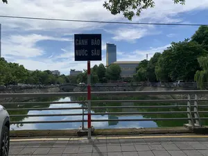 Trên biển báo màu đen có ghi dòng chữ màu trắng: 'CẢNH BÁO NƯỚC SÂU'. Đây là khung cảnh một con kênh hoặc hồ trong một khu vực đô thị. Ở phía trước, một biển báo được gắn trên cột sơn sọc đỏ trắng cảnh báo về vùng nước sâu. Một lan can bằng kim loại chạy dọc theo vỉa hè để đảm bảo an toàn. Bên kia kênh là một con đường với nhiều cây xanh và một số ô tô đang đỗ. Phía sau là các tòa nhà, bao gồm một tòa nhà cao tầng hiện đại bằng kính. Ở phía xa có thể thấy một cây cầu vòm bằng đá. Bầu trời trong xanh có mây trắng. Khung cảnh chung cho thấy một ngày nắng đẹp và yên tĩnh bên một con kênh trong thành phố.。（日本語訳）。黒い看板に「警告 水深注意」と白い文字で書かれています。これは都市部にある運河または湖の風景です。手前には、赤と白の縞模様に塗られた柱に、水深が深いことを警告する標識が取り付けられています。安全確保のため、歩道に沿って金属製の手すりが走っています。運河の向こう側は、緑の木々が多い道で、数台の車が駐車しています。後方には、近代的なガラス張りの高層ビルを含む建物があります。遠くには石造りのアーチ橋が見えます。空は青く澄んでおり、白い雲があります。全体的な風景は、都市の運河沿いの、晴れた穏やかな一日を示しています。