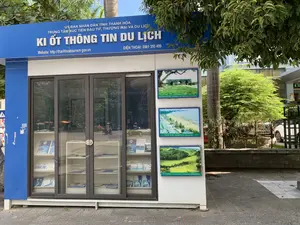 Bức ảnh chụp một ki-ốt thông tin du lịch màu xanh và trắng đặt trên vỉa hè, dưới bóng cây xanh. Trên biển hiệu lớn của ki-ốt có các dòng chữ: "ỦY BAN NHÂN DÂN TỈNH THANH HÓA", "TRUNG TÂM XÚC TIẾN ĐẦU TƯ, THƯƠNG MẠI VÀ DU LỊCH", và dòng chữ lớn nhất "KI ỐT THÔNG TIN DU LỊCH". Ngoài ra còn có thông tin website "http://thanhhoatourism.gov.vn" và số điện thoại "ĐIỆN THOẠI: 0961 355 499". Bên trong ki-ốt, qua cửa kính, là các kệ trưng bày nhiều tờ rơi và ấn phẩm du lịch. Phía bên hông ki-ốt có treo ba bức ảnh quảng bá các địa danh nổi tiếng của tỉnh Thanh Hóa, bao gồm: "THÀNH NHÀ HỒ (CITADEL OF THE HO DYNASTY)", "SẦM SƠN (SAM SON BEACH)", và "PÙ LUÔNG (PU LUONG NATURE RESERVE)". Hình ảnh phản chiếu trên cửa kính cho thấy cảnh đường phố với người và xe máy đang qua lại, trong đó có một người đội nón lá đang đi xe máy.。（日本語訳）。写真には、緑の木陰の歩道に設置された、青と白の観光案内キオスクが写っている。キオスクの大きな看板には、「THANH HOA省人民委員会」、「投資・貿易・観光促進センター」、そして最も大きな文字で「観光案内キオスク」と書かれている。さらに、ウェブサイト「http://thanhhoatourism.gov.vn」と電話番号「電話：0961 355 499」の情報もある。キオスクの側面には、タインホア省の有名な観光地を宣伝する3枚の写真が掲げられており、それには「胡朝の城塞（CITADEL OF THE HO DYNASTY）」、「サムソン（SAM SON BEACH）」、そして「プールオン（PU LUONG NATURE RESERVE）」が含まれる。ガラス窓の反射には、人々やバイクが行き交う通りの様子が映っており、その中にはノンラーをかぶってバイクに乗っている人もいる。
