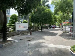 Bức ảnh chụp một con phố tại thành phố Thanh Hóa vào một ngày nắng. Cảnh vật cho thấy một vỉa hè rộng rãi được lát gạch, hai bên trồng nhiều cây xanh tạo bóng mát. Ở bên trái, có một tấm biển quảng cáo lớn màu trắng của Ngân hàng Hợp tác xã Việt Nam (Co-opBank). Trên biển có ghi rõ thông tin: "Ngân hàng Hợp tác xã Việt Nam", logo "Co-opBank", và địa chỉ "Tầng 3 Tòa nhà Khách sạn Sao Mai - Số 20 Phan Chu Trình - P. Điện Biên - TP. Thanh Hóa". Trên vỉa hè có một con chó nhỏ màu nâu đang ngồi. Phía xa là lòng đường với một vài chiếc ô tô đang đỗ và di chuyển. Khung cảnh tổng thể khá yên tĩnh, không có nhiều người qua lại.。（日本語訳）。写真は、晴れた日のタインホア市のある通りを撮影したものです。風景は、タイルで舗装された広い歩道を示しており、両側には日陰を作るために多くの緑の木が植えられています。左側には、ベトナム協同組合銀行（Co-opBank）の大きな白い看板があります。看板には、「ベトナム協同組合銀行」、ロゴ「Co-opBank」、および住所「タインホア市ディエンビエン区ファン・チュー・チン通り20番地 サオマイホテルビル3階」という情報が明記されています。歩道には、茶色い小さな犬が座っています。遠くには車道があり、数台の車が駐車したり移動したりしています。全体の風景はかなり静かで、行き交う人は多くありません。