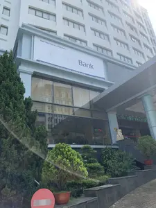 Bức ảnh chụp mặt tiền của một tòa nhà cao tầng hiện đại. Trên tòa nhà có một biển hiệu lớn ghi 'Ngân hàng Hợp tác xã Việt Nam' và 'Co-op Bank'. Dòng chữ nhỏ hơn trên biển hiệu cung cấp thông tin địa chỉ: 'Tầng 3 Tòa nhà Khách sạn Sao Mai - Số 20 Phan Chu Trinh - P. Điện Biên - TP Thanh Hóa' và trang web 'www.co-opbank.vn'. Tòa nhà này là Khách sạn Sao Mai. Phía trước tòa nhà có nhiều cây xanh và cây cảnh. Ở tiền cảnh bên trái có một biển báo giao thông 'Cấm đi ngược chiều'. Qua các ô cửa kính lớn ở tầng trệt, có thể nhìn thấy một phần nội thất bên trong, bao gồm một biển hiệu đèn ghi 'Bar Cafe'. Gần lối vào có một bức tượng sư tử đá màu trắng và một bảng điện tử chữ đỏ nhưng không đọc rõ nội dung. Trên cửa kính có chữ 'SAO MAI HOTEL' và 'ĐẨY VÀO'. Không có hoạt động của con người nào được ghi lại rõ ràng trong ảnh.。（日本語訳）。写真は近代的な高層ビルの正面を撮影したものです。ビルには「Ngân hàng Hợp tác xã Việt Nam」（ベトナム協同組合銀行）と「Co-op Bank」と書かれた大きな看板があります。看板の小さな文字には、住所「Tầng 3 Tòa nhà Khách sạn Sao Mai - Số 20 Phan Chu Trinh - P. Điện Biên - TP Thanh Hóa」（サオマイホテルビル3階 - ファン・チュー・チン通り20番 - ディエンビエン区 - タインホア市）とウェブサイト「www.co-opbank.vn」の情報が記載されています。このビルはサオマイホテルです。ビルの前には多くの緑の木々や観葉植物があります。前景の左側には「Cấm đi ngược chiều」（一方通行）の交通標識があります。1階の大きなガラス窓を通して、「Bar Cafe」と書かれた電光看板を含む、内部のインテリアの一部が見えます。入口の近くには白い石の獅子の像と、内容ははっきりと読み取れませんが赤い文字の電光掲示板があります。ガラスのドアには「SAO MAI HOTEL」と「ĐẨY VÀO」（押す）という文字があります。写真には人間の活動ははっきりと写っていません。