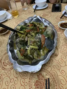 Hình ảnh chụp cận cảnh một bàn ăn, có thể là ở một nhà hàng hoặc tại gia đình. Tâm điểm của bức ảnh là một đĩa cá om dưa lớn đặt trên một chiếc đĩa kim loại hình con cá. Món ăn bao gồm một con cá nguyên con được nấu cùng với dưa cải muối, cà chua và các loại rau thơm. Trên bàn ăn được trải một tấm khăn trải bàn màu vàng có hoa văn. Xung quanh đĩa cá là các vật dụng ăn uống như bát sứ trắng, đũa, chén nước chấm, một ly bia đang uống dở và một ly nước lọc. Một đôi đũa đang gắp thức ăn từ đĩa cá, cho thấy mọi người đang trong một bữa ăn.。（日本語訳）。食卓のクローズアップ写真で、レストランか家庭での食事と思われる。写真の中心は、魚の形をした金属製の皿に盛られた大きな「カーオムズア」（魚と高菜の煮込み）の皿である。この料理は、魚一匹丸ごとを高菜の漬物、トマト、各種香草と一緒に煮込んだものである。食卓には模様の入った黄色いテーブルクロスが敷かれている。一膳の箸が魚の皿から食べ物を取っており、人々が食事中であることを示している。