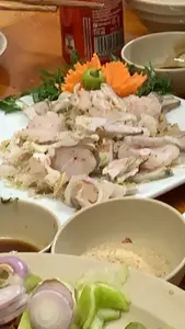 Đây là hình ảnh một bàn ăn, có thể là trong một nhà hàng hoặc tại gia. Món ăn chính trên đĩa sứ trắng là một món gỏi, có vẻ là gỏi cá, bao gồm những lát cá mỏng trộn với các loại gia vị. Món ăn được trang trí bằng cà rốt tỉa hình hoa và rau thơm. Ở phía trước, có các bát nhỏ đựng đồ ăn kèm và gia vị, bao gồm một bát đựng gia vị dạng bột (có thể là thính hoặc muối tiêu) và một đĩa có hành tím thái lát. Ở phía sau, có một lon nước ngọt Coca-Cola màu đỏ.。（日本語訳）。これは食卓の写真で、レストランか自宅かもしれません。白い磁器の皿の上にあるメイン料理は和え物（ゴイ）で、魚の和え物（ゴイ・カー）のようです。薄切りの魚を各種スパイスで混ぜ合わせたものです。料理は花形に飾り切りされた人参と香草で飾られています。手前には、付け合わせや調味料を入れるための小さな器があり、粉末状の調味料（おそらくティンまたは塩コショウ）を入れた器と、スライスした赤玉ねぎの皿が含まれています。後ろには、赤いコカ・コーラの缶があります。