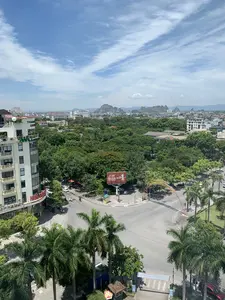 Bức ảnh chụp một góc nhìn toàn cảnh từ trên cao của một thành phố, có thể là thành phố Thanh Hóa, vào một ngày nắng đẹp. Khung cảnh cho thấy một ngã tư hoặc vòng xuyến với nhiều cây xanh và các tòa nhà bao quanh.。**Chi tiết văn bản trên các biển hiệu:**。*   Trên tòa nhà cao tầng màu trắng bên trái: Phía trên cùng có chữ "THANH HOA GROUP". Phía dưới có biển hiệu màu đỏ với chữ "NHA SACH" (Nhà Sách).。*   Trên tấm biển quảng cáo lớn (billboard) ở giữa ngã tư: Có logo và chữ "GENERALI", cùng với dòng chữ "TỰA VỮNG TÀI CHÍNH - BẢO HIỂM SỐ 1 Ý" và hashtag "#SốngNhưÝ".。*   Trên một tòa nhà khác ở phía xa: Có thể thấy chữ "DAI VIET PALACE".。*   Trên cổng chào trang trí màu đỏ và vàng bắc qua đường: Có chữ nhưng không thể đọc rõ do khoảng cách và góc chụp.。**Mô tả cảnh quan và hoạt động:**。*   **Loại địa điểm:** Đây là một khu vực đô thị với đường phố, ngã tư, công viên và các tòa nhà cao tầng.。*   **Hoạt động:** Giao thông đô thị đang diễn ra với một vài chiếc ô tô và xe máy di chuyển trên đường. Đây là một cảnh sinh hoạt thường ngày trong thành phố.。*   **Các vật thể trong ảnh:**。*   **Công trình:** Nhiều tòa nhà cao tầng và nhà ở, một ngã tư đường phố, một cổng chào trang trí, và một biển quảng cáo lớn.。*   **Thiên nhiên:** Thành phố có rất nhiều cây xanh, bao gồm cả hàng cây cọ dọc theo con đường và cây phượng có hoa đỏ. Ở phía xa là những dãy núi đá vôi đặc trưng. Bầu trời trong xanh, có mây trắng.。*   **Phương tiện:** Có ô tô và xe máy đang lưu thông trên đường.。（日本語訳）。写真は、ある晴れた日に、上空から撮影されたある都市、おそらくタインホア市のパノラマビューを捉えています。**看板の文字詳細：**。*   左側の白い高層ビル：最上部に「THANH HOA GROUP」の文字。その下には「NHA SACH」（ニャーサック、書店）と書かれた赤い看板があります。*   交差点の中央にある大きな広告看板（ビルボード）：「GENERALI」のロゴと文字、そして「TỰA VỮNG TÀI CHÍNH - BẢO HIỂM SỐ 1 Ý」（安定した財政基盤 - イタリアNo.1保険）という文言とハッシュタグ「#SốngNhưÝ」（ソンニューイー、思い通りに生きる）があります。*   遠くにある別の建物：「DAI VIET PALACE」の文字が見えます。**風景と活動の描写：**。*   **場所の種類：**ここは道路、交差点、公園、高層ビルがある都市部です。*   **活動：**都市の交通が行われており、数台の自動車とバイクが道路を移動しています。これは都市の日常的な生活風景です。*   **写真の中の物体：**。*   **建造物：**多くの高層ビルと住宅、一つの道路の交差点、一つの装飾的な歓迎ゲート、そして一つの大きな広告看板。*   **自然：**都市には多くの緑樹があり、道に沿ったヤシの木の並木や赤い花をつけたホウオウボクを含みます。遠くには特徴的な石灰岩の山脈があります。空は青く澄み、白い雲があります。*   **乗り物：**自動車とバイクが道路を通行しています。