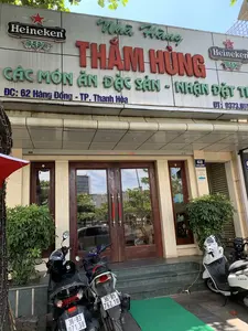 Bức ảnh chụp mặt tiền của một nhà hàng tên là 'Nhà Hàng Thăm Hùng' tại thành phố Thanh Hóa. Trên biển hiệu lớn có các dòng chữ: 'Nhà Hàng THĂM HÙNG', 'CÁC MÓN ĂN ĐẶC SẢN - NHẬN ĐẶT TIỆC'. Địa chỉ được ghi rõ là 'ĐC: 62 Hàng Đồng - TP. Thanh Hóa' và một phần số điện thoại 'ĐT: 0373.851...'. Hai bên biển hiệu chính có logo của thương hiệu bia Heineken. Trên cột nhà có biển số nhà '62 HÀNG ĐỒNG' và một tấm biển nhỏ với nội dung 'NÓI LỜI HAY LÀM VIỆC TỐT HÀNH ĐỘNG THÂN THIỆN'. Phía trước nhà hàng có một vài chiếc xe máy đang đỗ, trong đó có một chiếc xe máy điện màu trắng đang được cắm sạc. Lối vào là cửa kính khung gỗ, hai bên có trồng cây cảnh.。（日本語訳）。タインホア市にある「Nhà Hàng Thăm Hùng（タムフンレストラン）」という名前のレストランの正面を撮影した写真。大きな看板には「Nhà Hàng THĂM HÙNG」、「CÁC MÓN ĂN ĐẶC SẢN - NHẬN ĐẶT TIỆC（特産料理 - 宴会予約受付）」という文字がある。住所は「ĐC: 62 Hàng Đồng - TP. Thanh Hóa（住所：タインホア市ハン・ドン通り62番地）」と明記されており、電話番号の一部「ĐT: 0373.851...」も記載されている。メイン看板の両側には、ハイネケンビールのブランドロゴがある。建物の柱には「62 HÀNG ĐỒNG」という番地表示板と、「NÓI LỜI HAY LÀM VIỆC TỐT HÀNH ĐỘNG THÂN THIỆN（良い言葉を話し、良い行いをし、親しみやすく行動する）」という内容の小さな看板がある。レストランの前には数台のバイクが停まっており、その中には充電中の白い電動バイクが1台ある。入り口は木枠のガラス扉で、両脇には観葉植物が植えられている。
